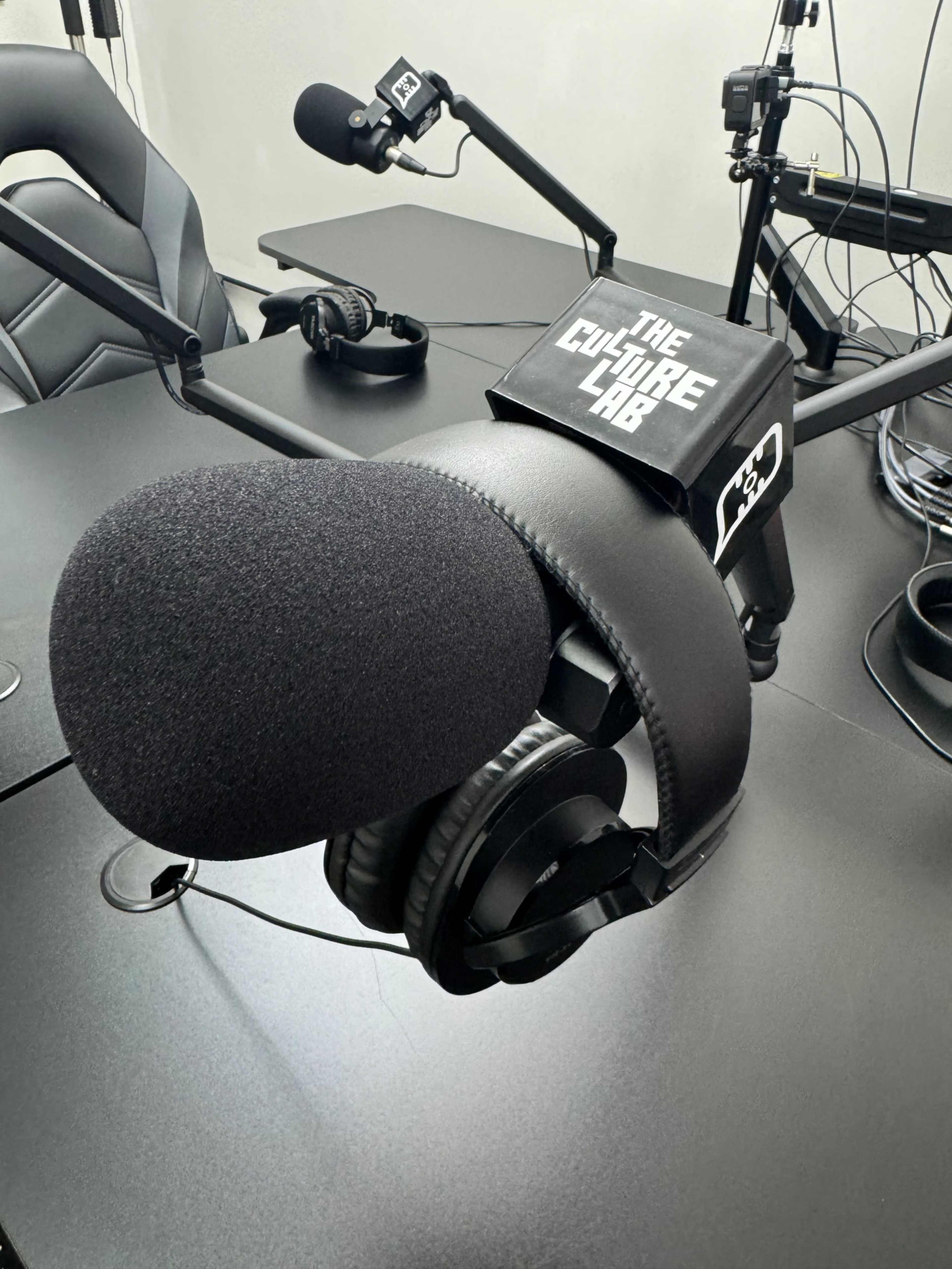 The image shows a microphone mounted on an adjustable arm with headphones resting on it, positioned on a desk in a recording studio.