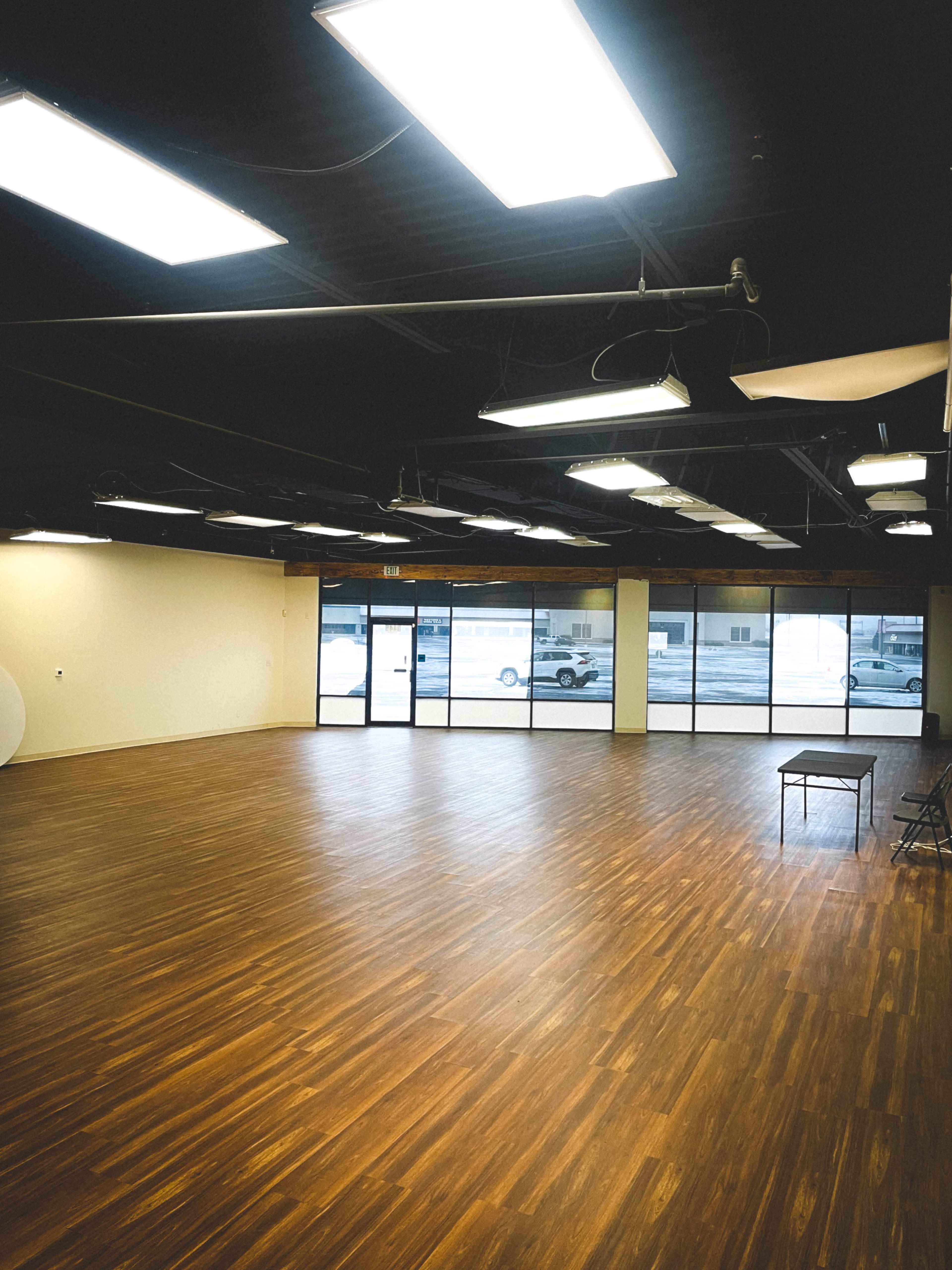 The image shows a spacious, empty room with wooden flooring and large windows allowing light to enter.