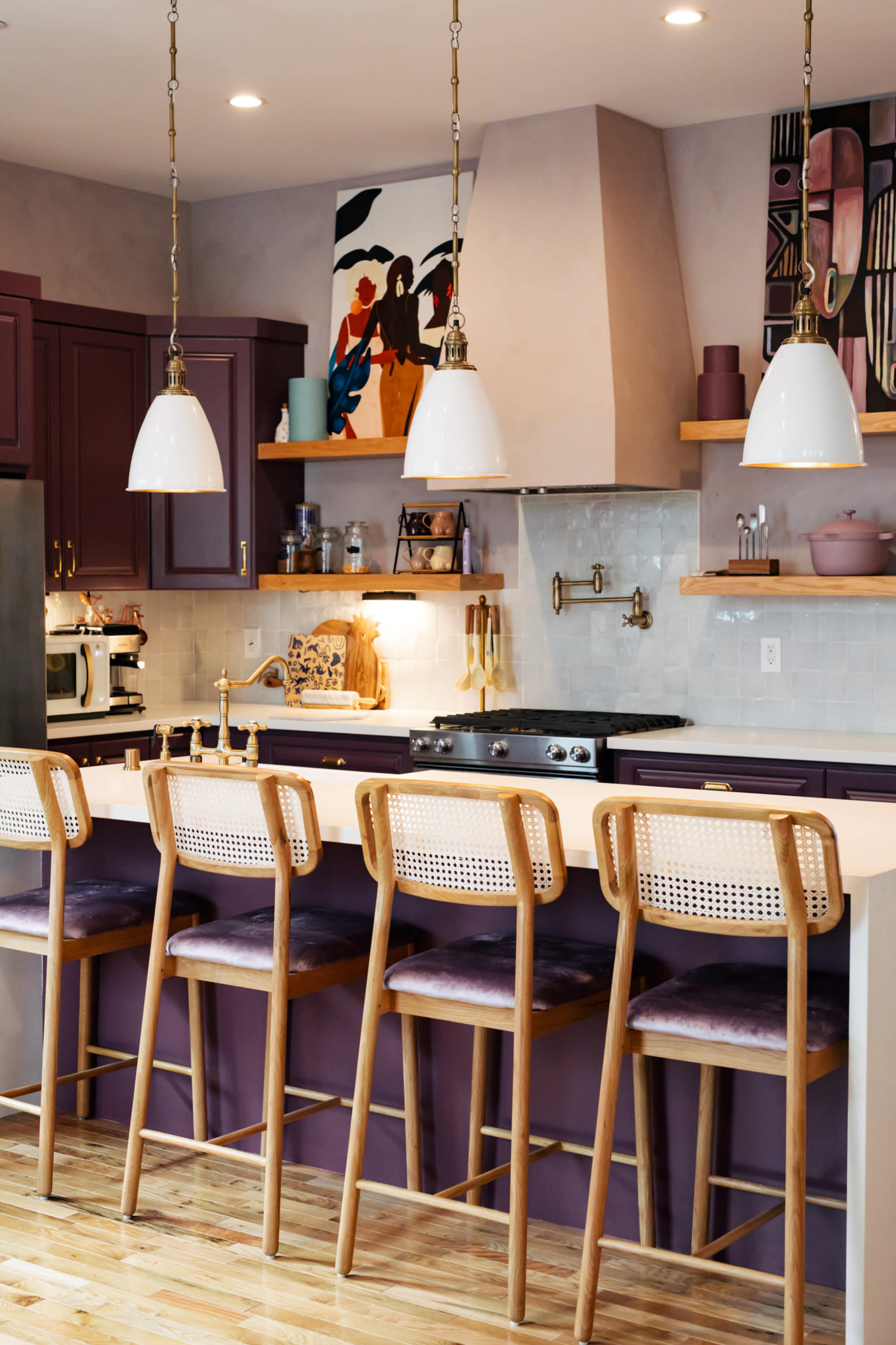 A modern kitchen features a bar with four wooden stools, pendant lighting, and purple cabinetry, alongside a stove and artistic wall decor.