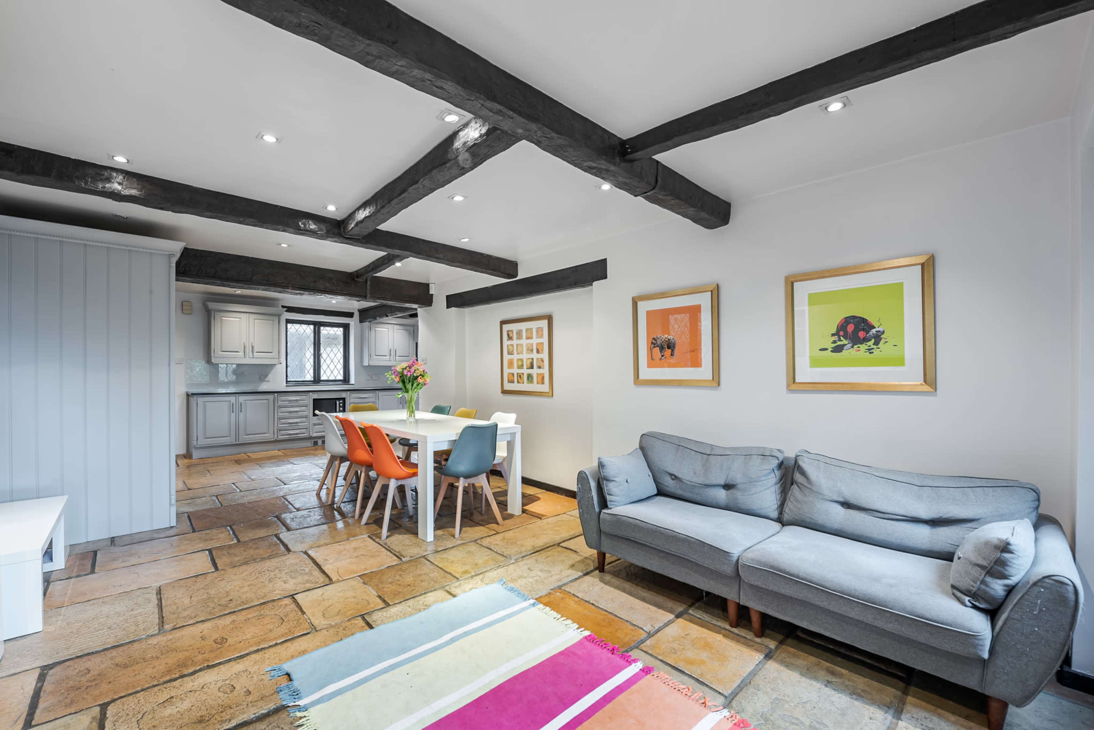 The image shows a bright, open living space with a gray sofa, a dining table with colorful chairs, and exposed wooden beams in the ceiling.