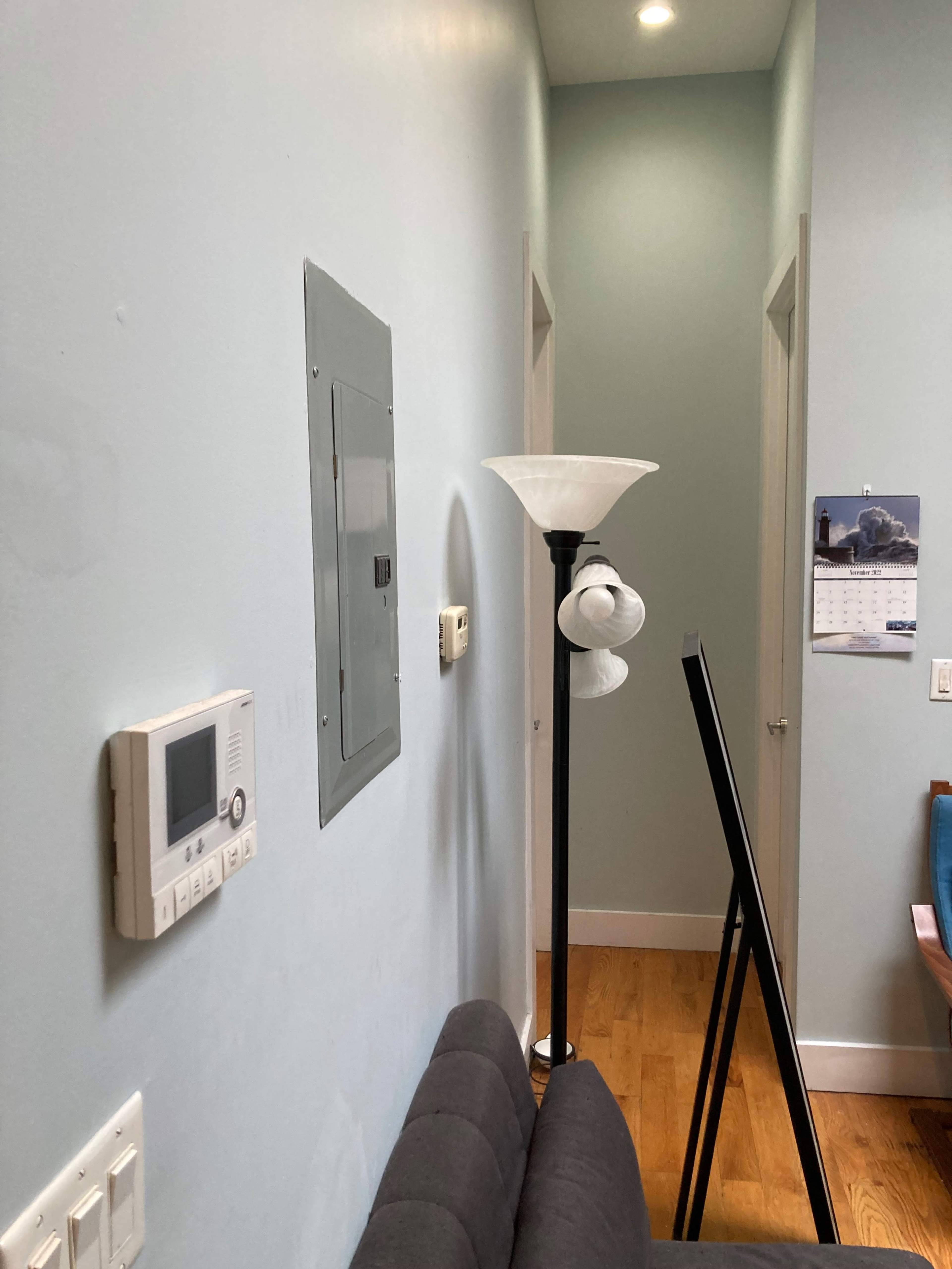 A light blue wall with a light switch, a thermostat, a lamp, and a calendar, along with two doorways in a hallway.