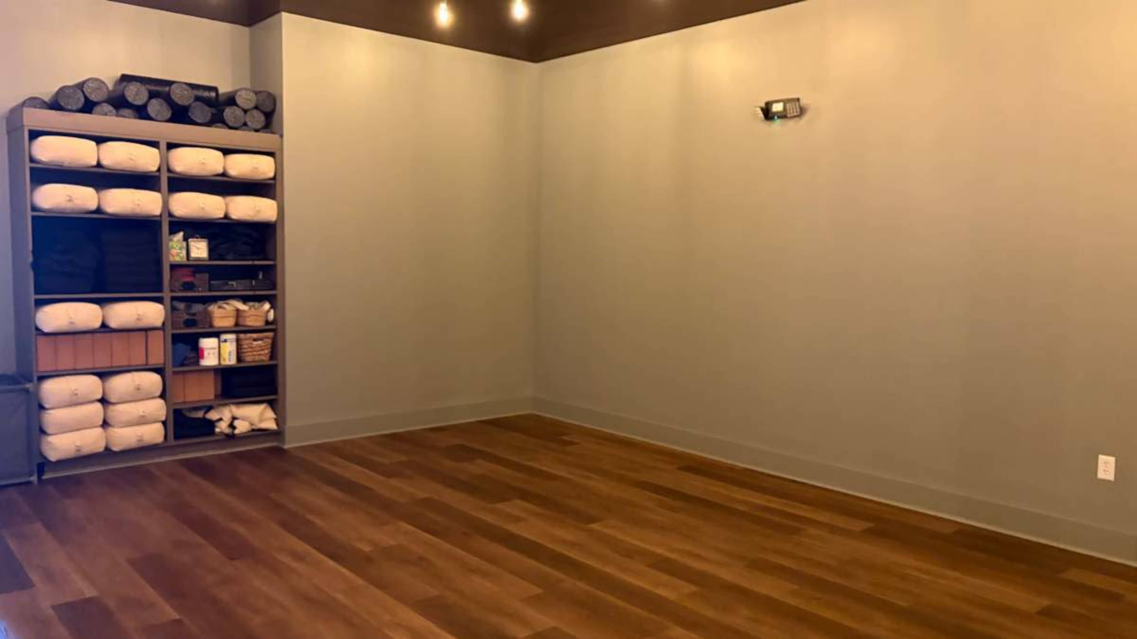 The image shows a spacious room with wooden flooring, a light-colored wall, and shelves containing neatly arranged yoga mats and towels.