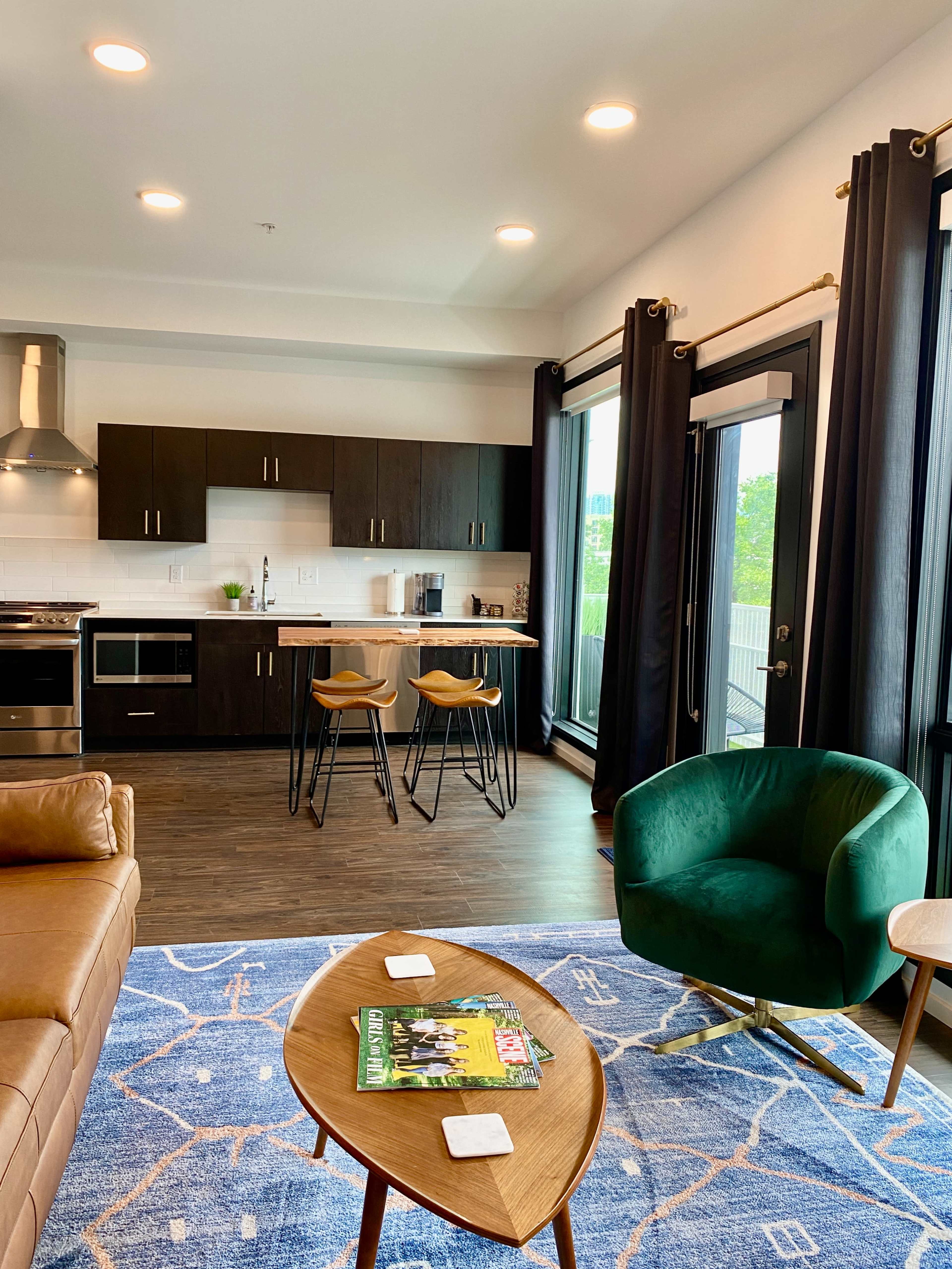 A modern kitchen and living area features dark wooden cabinets, a kitchen island with stools, and large windows with black curtains.