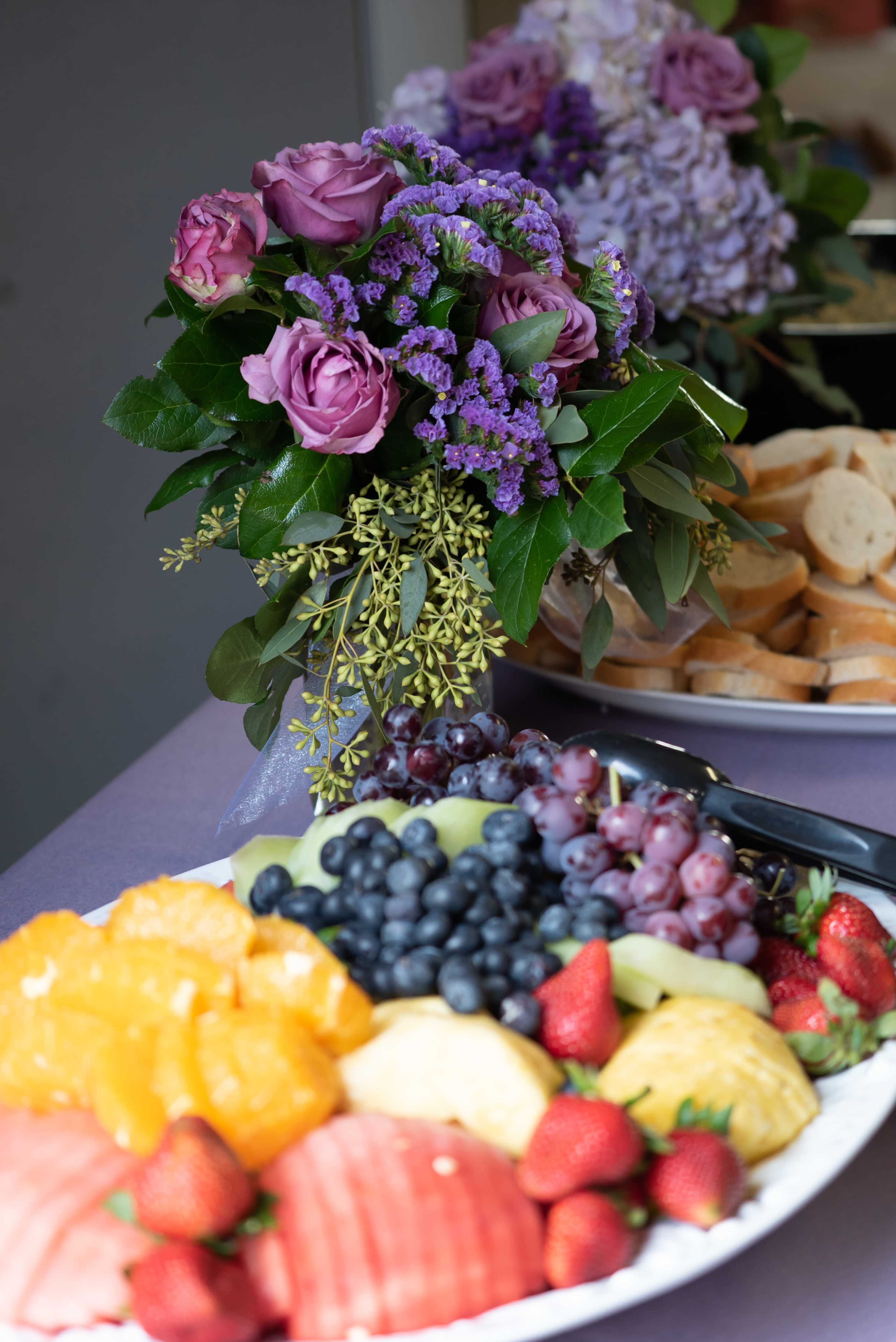 A platter of assorted fruits, including watermelon, strawberries, and blueberries, is displayed next to a vase of purple and green flowers.