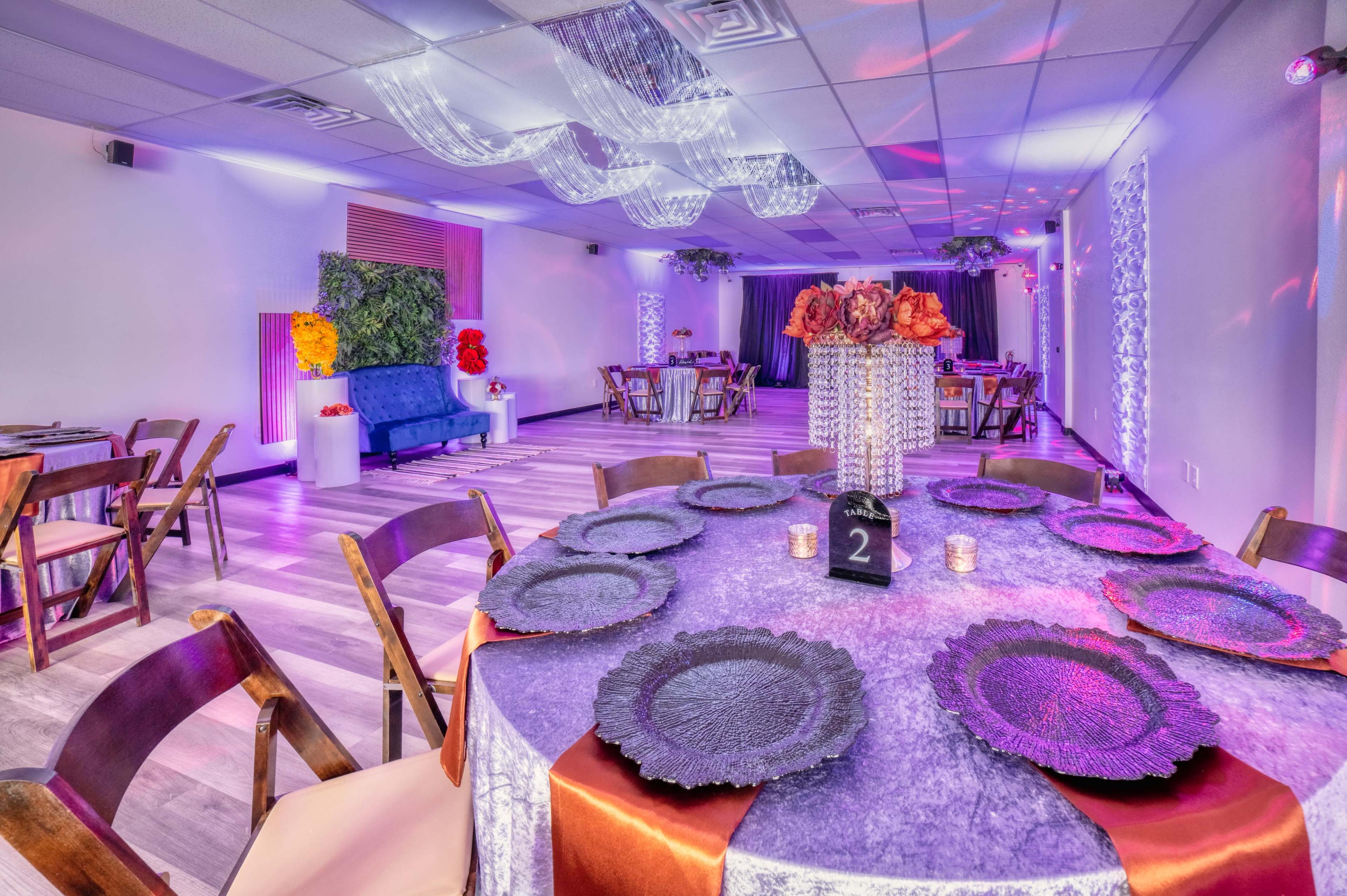 A beautifully arranged event space features round tables with decorative tableware, a blue couch, and floral centerpieces under colorful lighting.