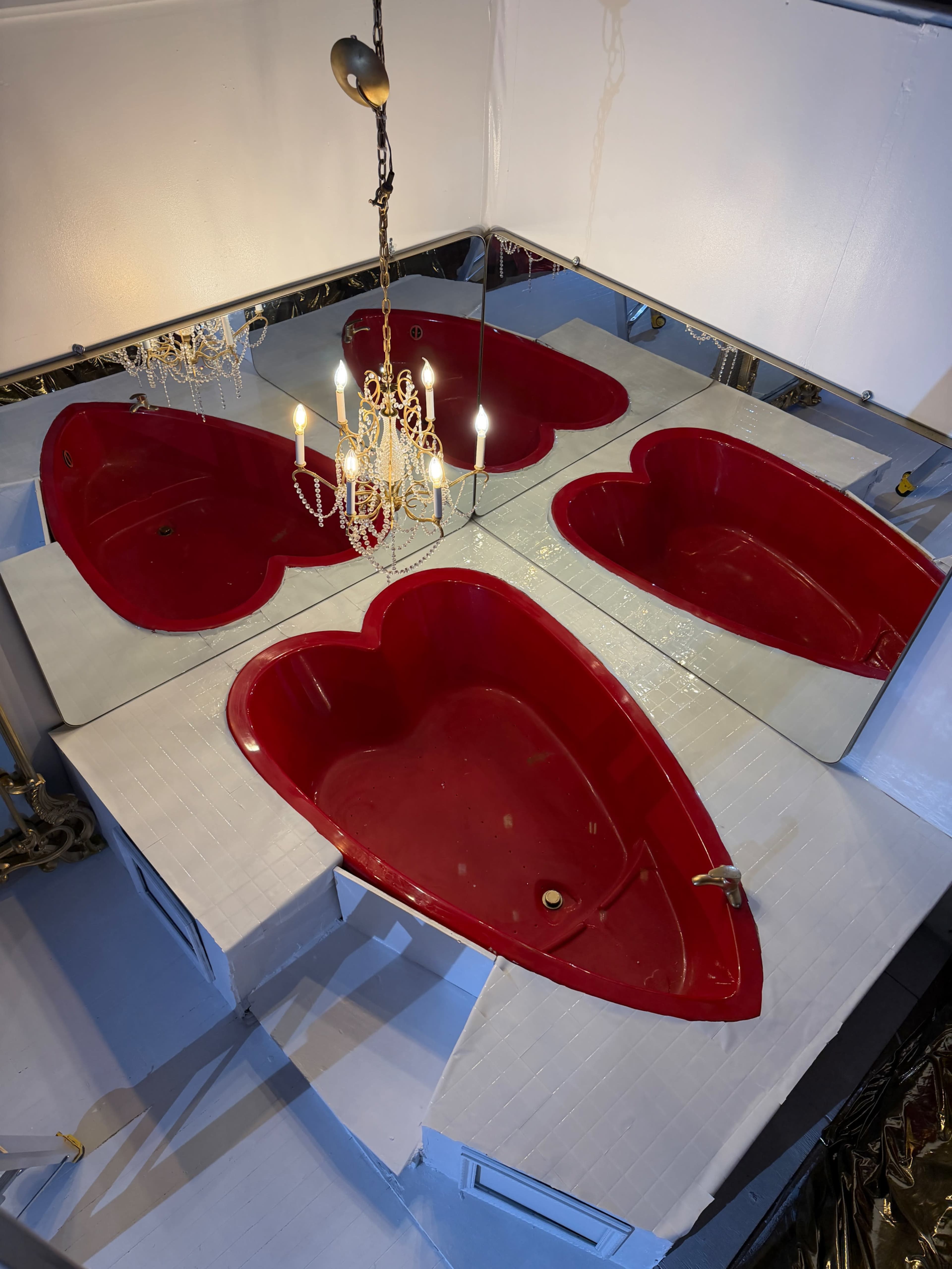 The image shows a room featuring four heart-shaped red bathtubs arranged under a chandelier, reflecting in surrounding mirrors.