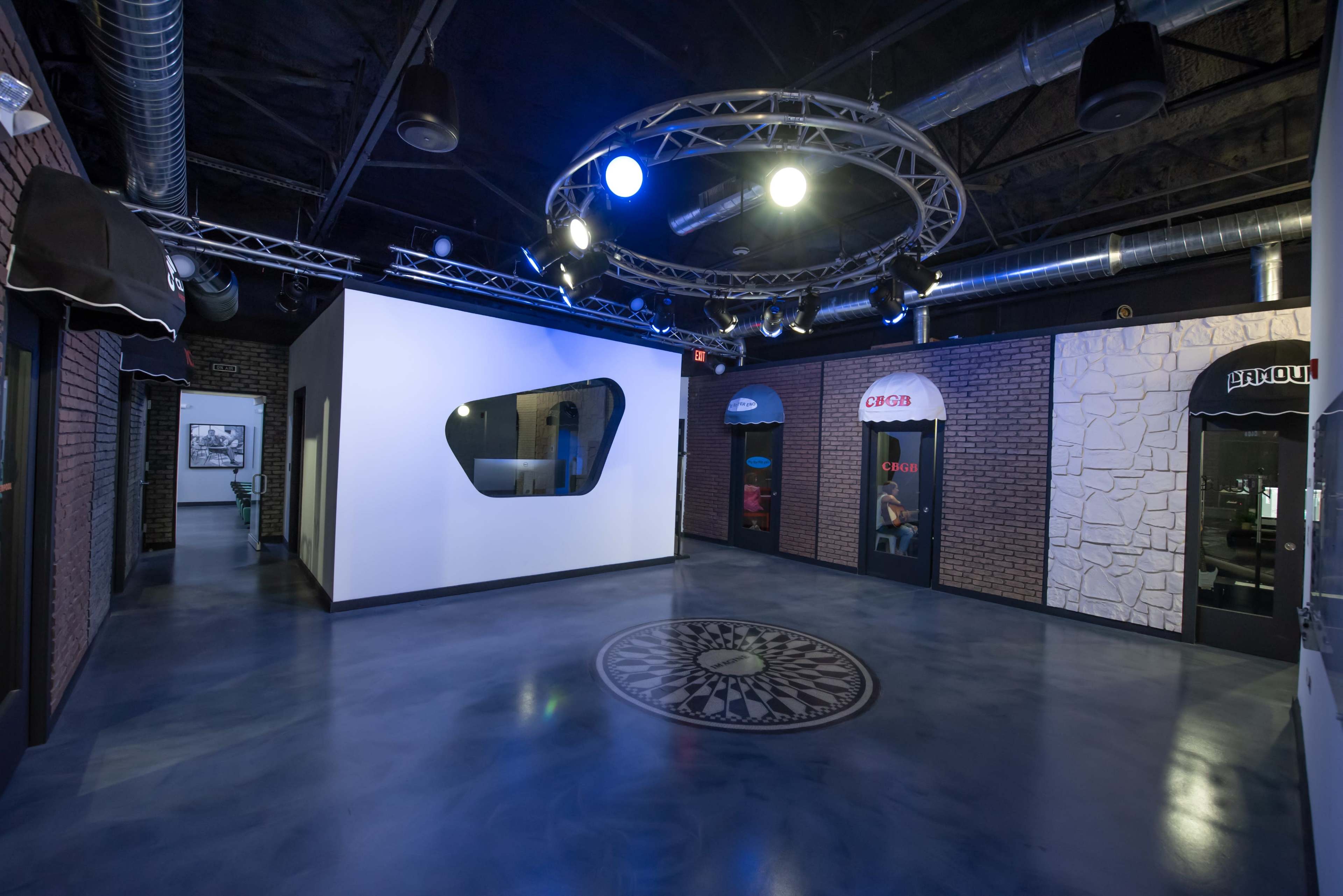 A modern indoor space with a circular design feature on the floor, illuminated by overhead lights, and flanked by doorways resembling storefronts.