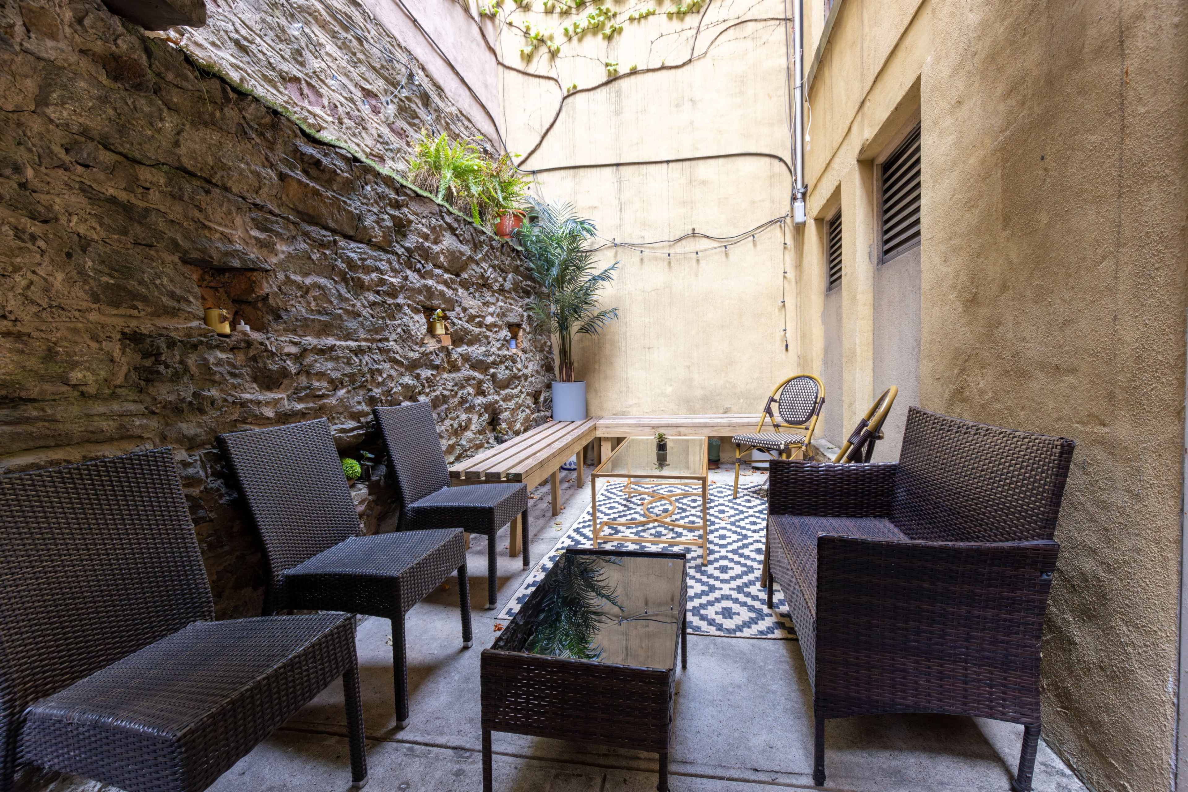 The image shows a narrow outdoor patio area with a stone wall, several wicker chairs, a glass coffee table, and potted plants.