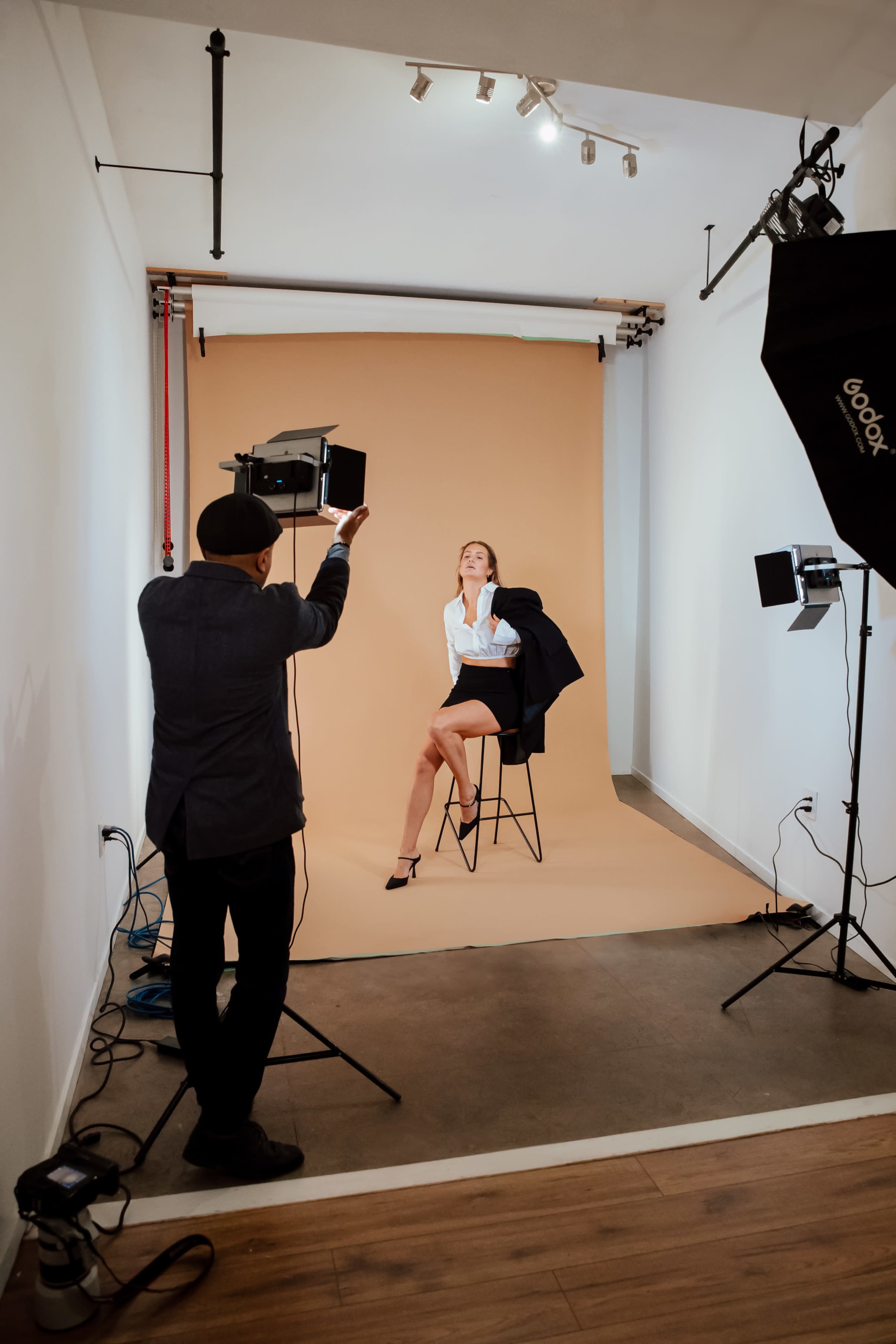 A photographer captures a model posing on a stool in a studio with a tan backdrop and professional lighting equipment.