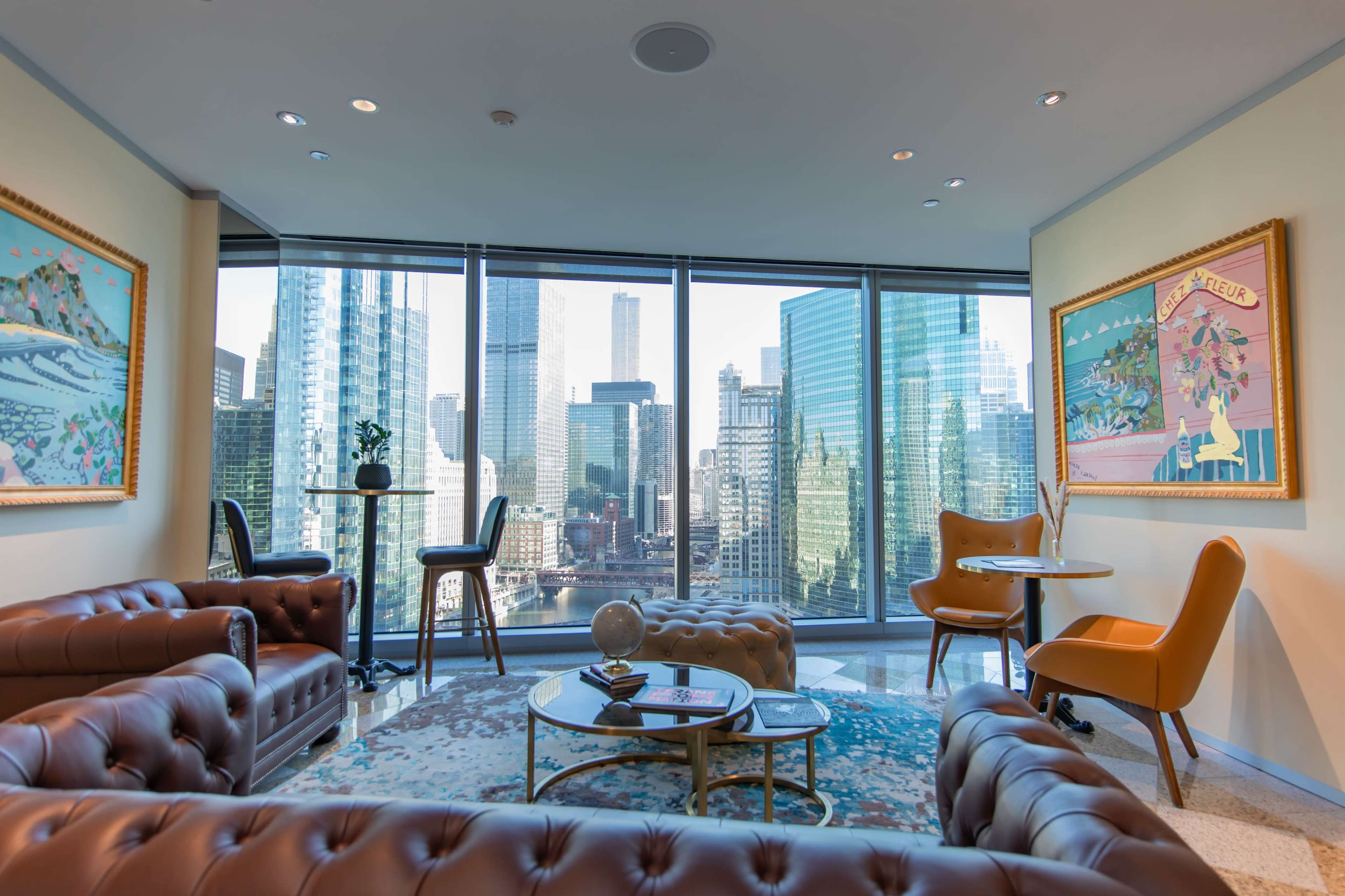 The image shows a modern lounge area with brown leather sofas, a glass coffee table, and large windows showcasing a view of skyscrapers in a cityscape.