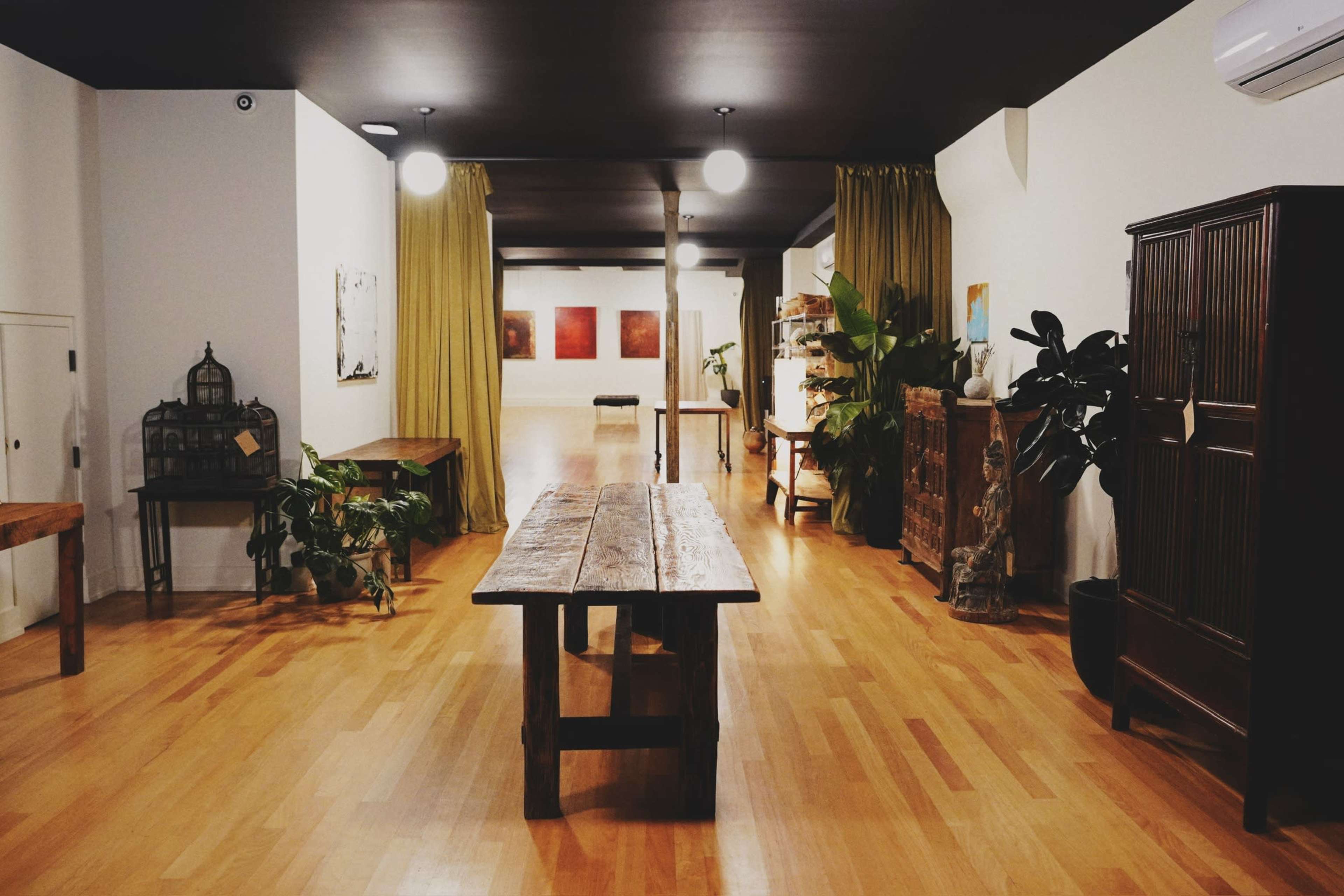 The image shows a spacious, well-lit interior featuring wooden flooring, a long wooden table in the center, and various plants and furniture arrangements along the walls.