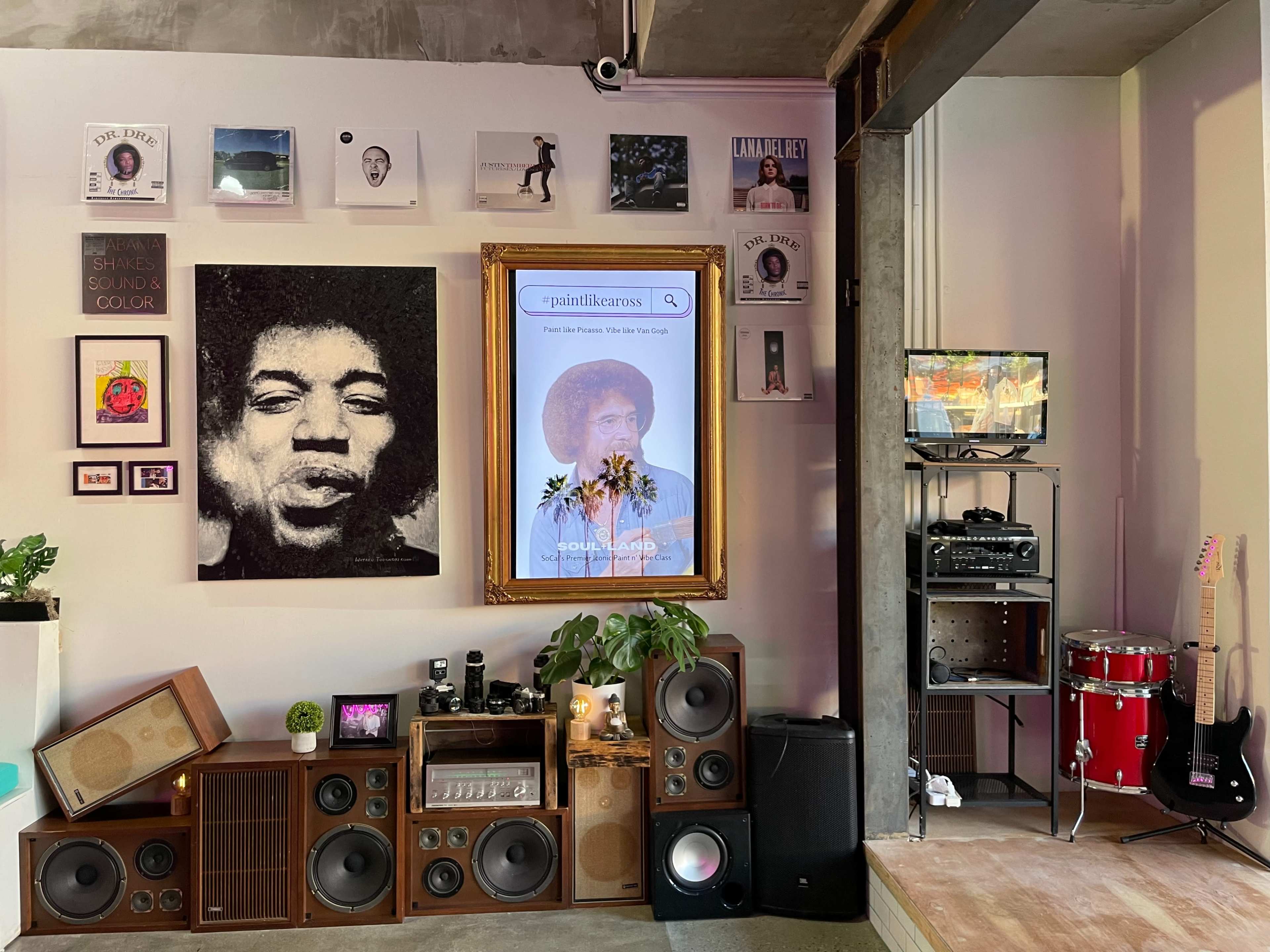 A wall featuring a variety of framed art pieces, a central portrait of Jimi Hendrix, a vintage stereo setup, and a small television beside a drum set.