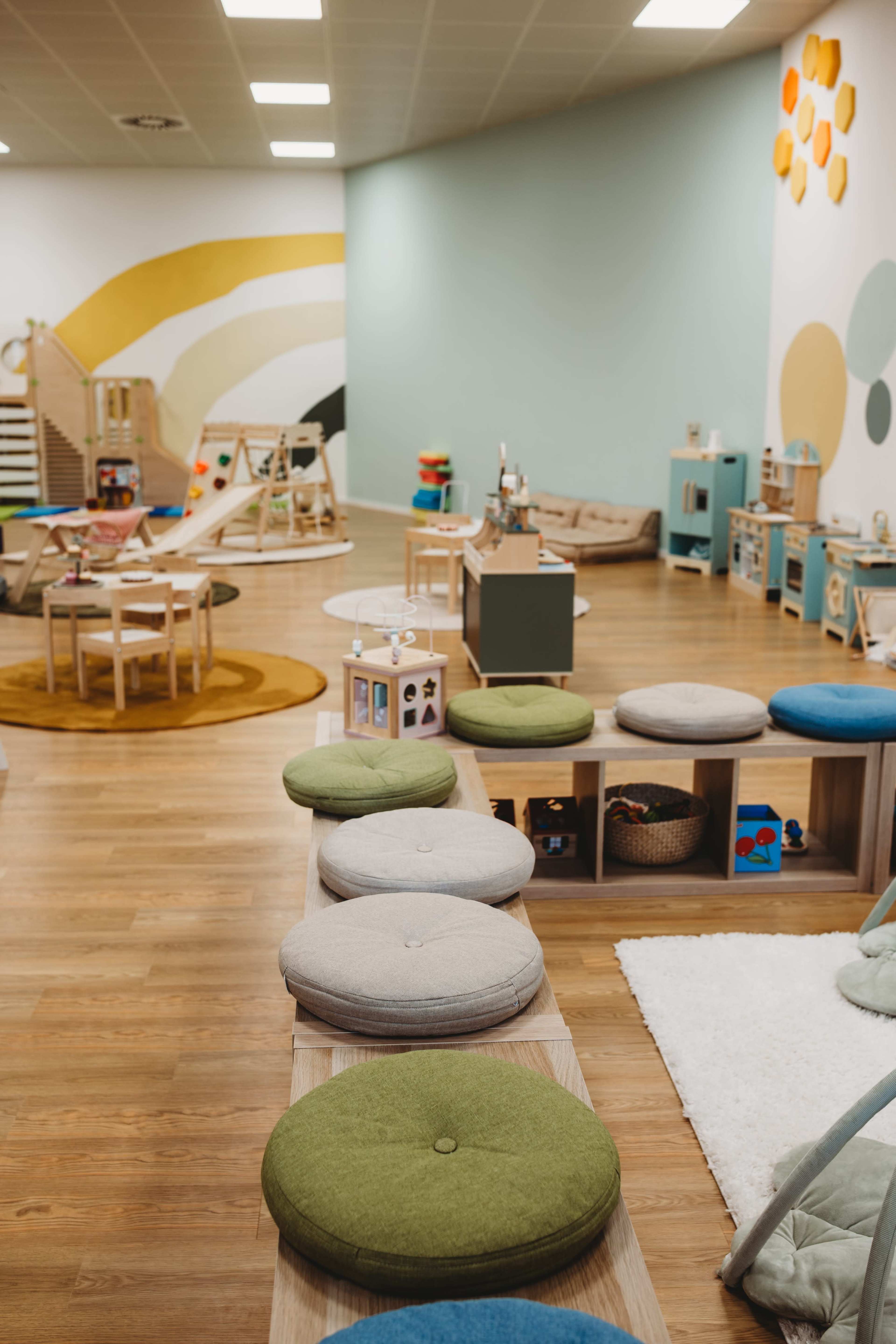 The image shows a colorful children's playroom filled with various seating options, play equipment, and educational toys arranged neatly on shelves.