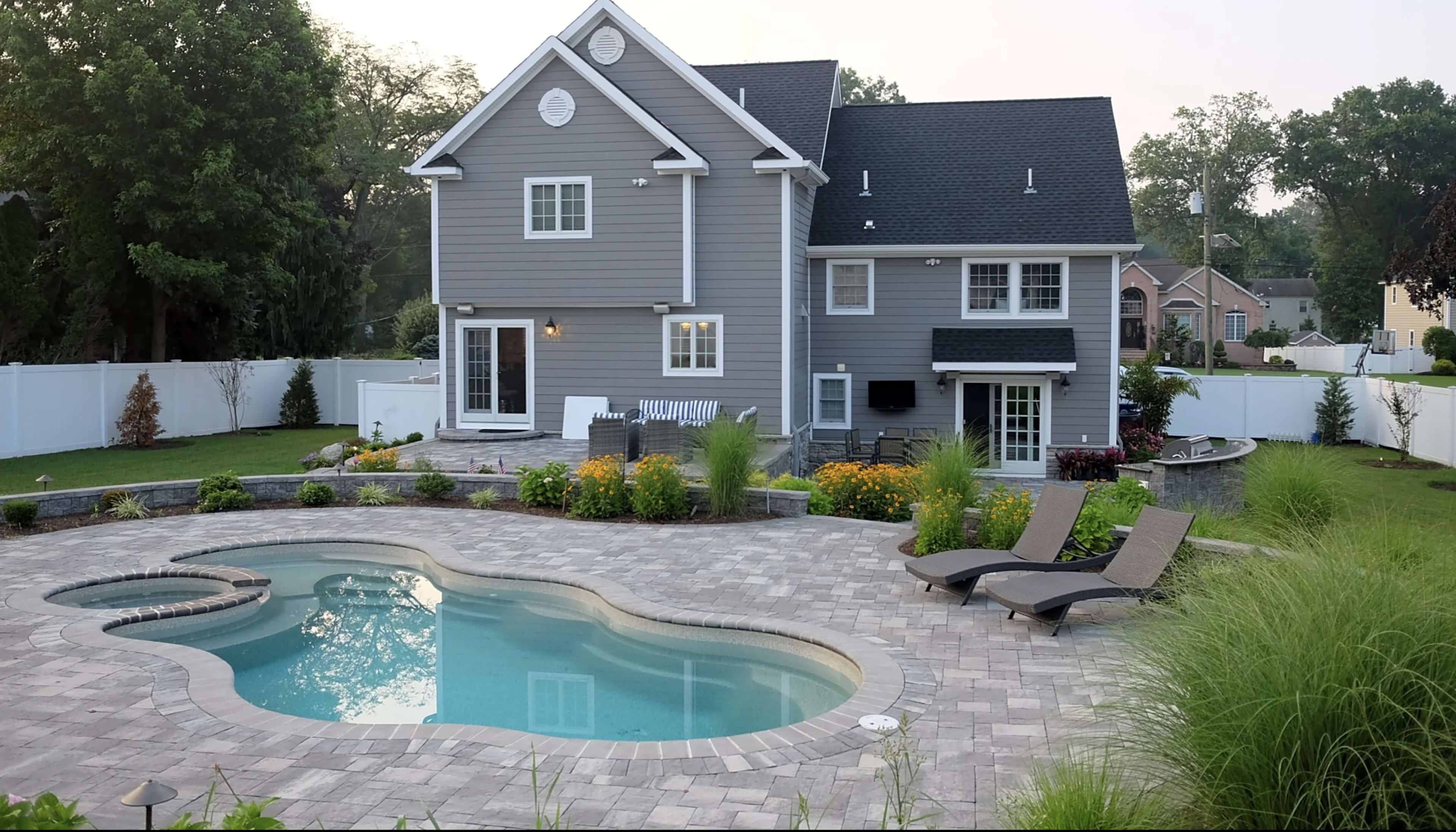 A spacious gray house with a landscaped yard features a swimming pool surrounded by lounge chairs and flower beds.