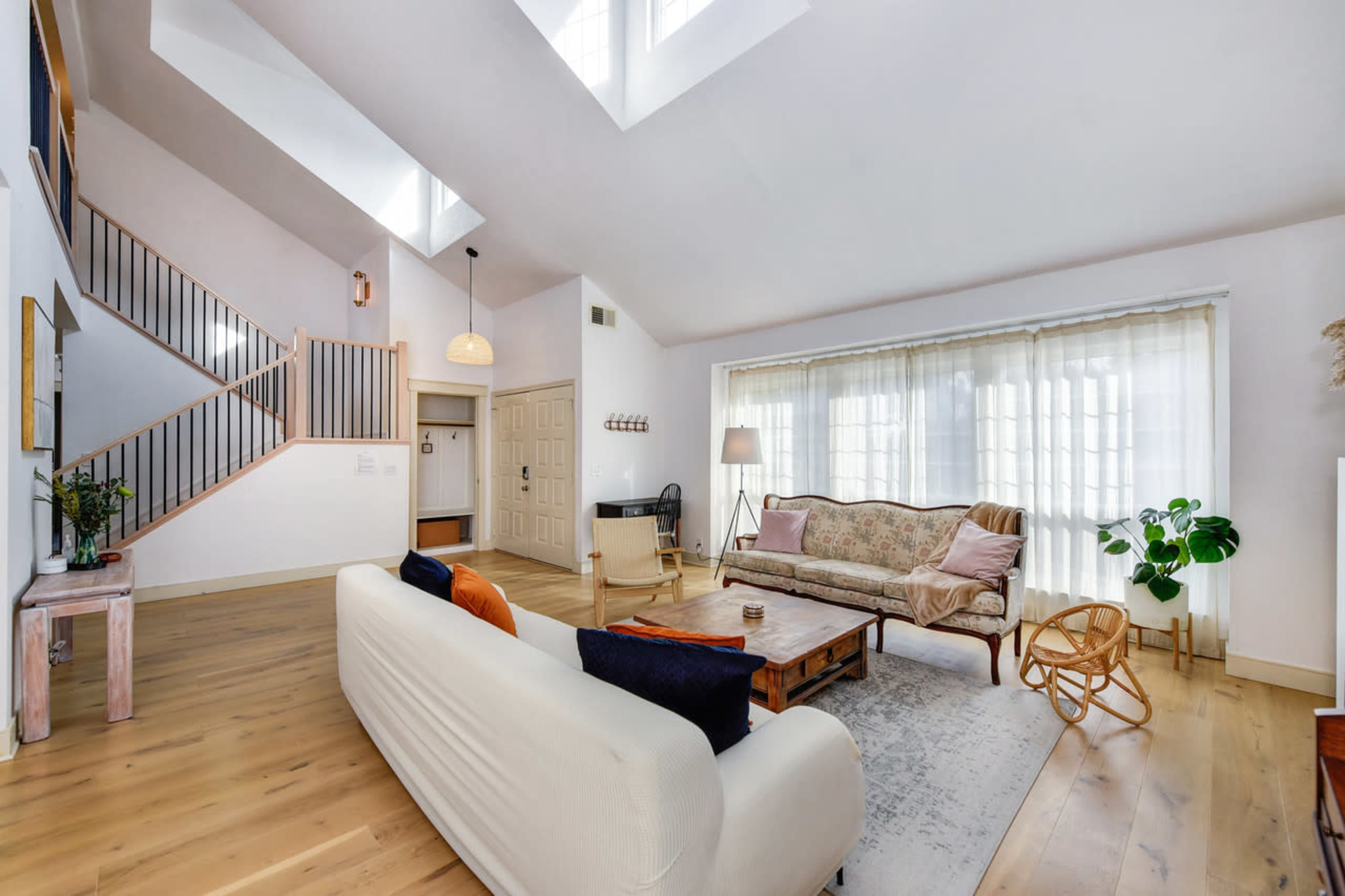 A spacious living room with a high ceiling, featuring a sofa, armchair, coffee table, and large windows letting in natural light.