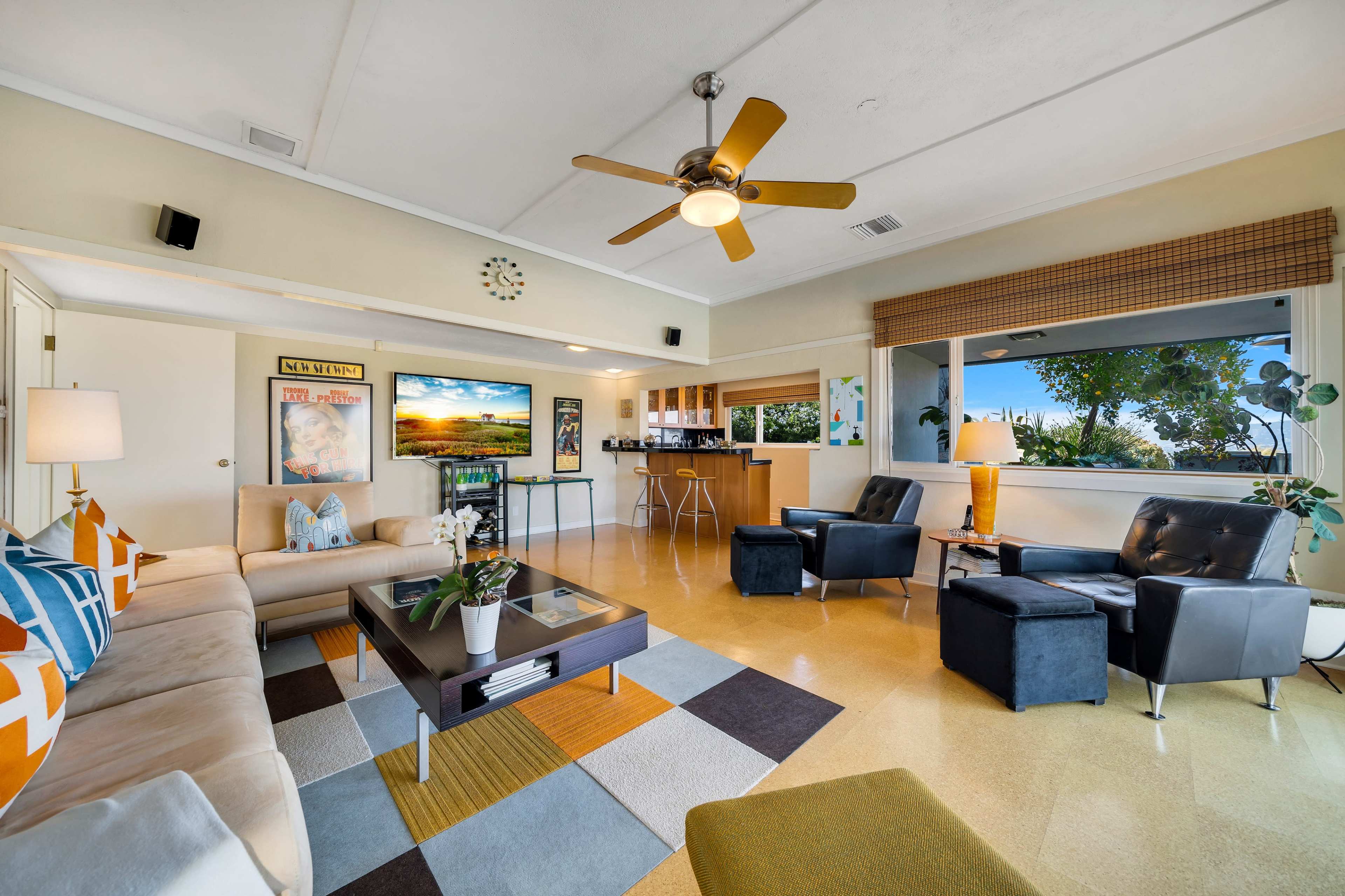 A spacious living room features a light-colored couch, two dark armchairs, a coffee table with decor, and a dining area visible in the background, all under a ceiling fan.