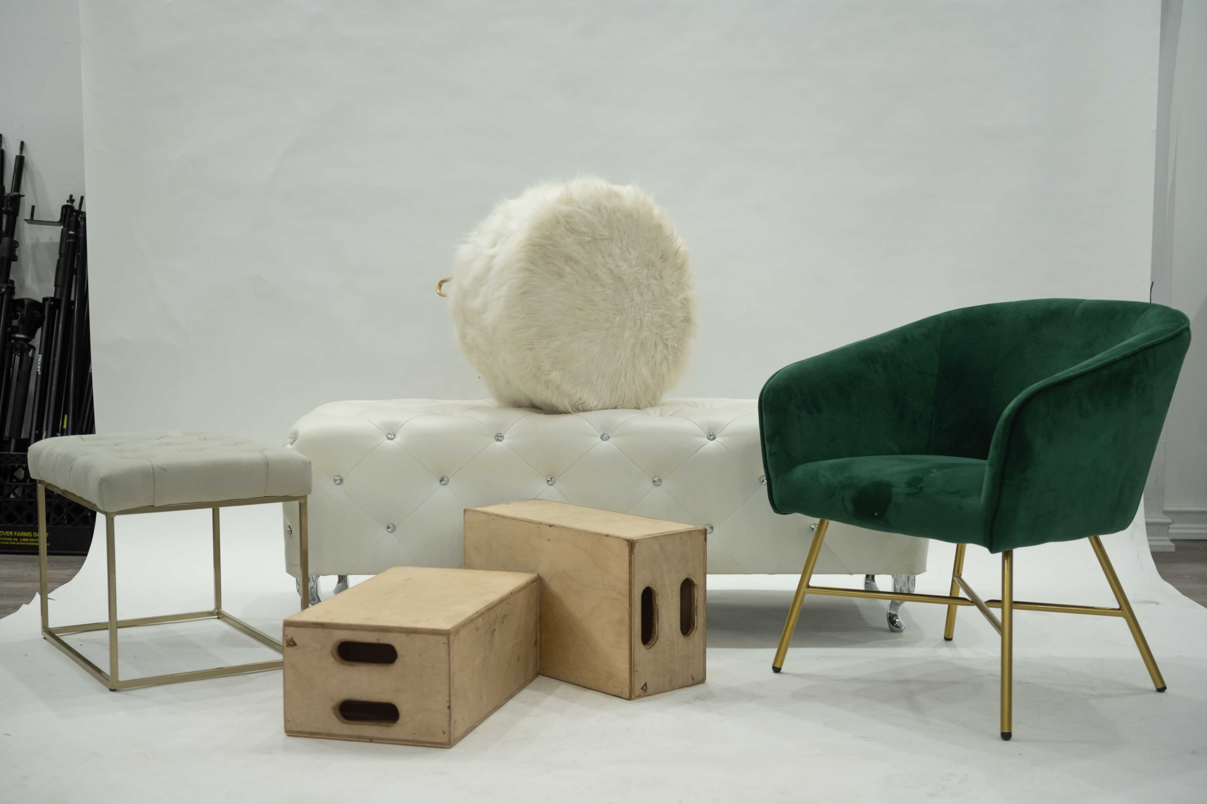 A green upholstered chair, a tufted white bench, two wooden boxes, and a round faux fur cushion against a plain backdrop.