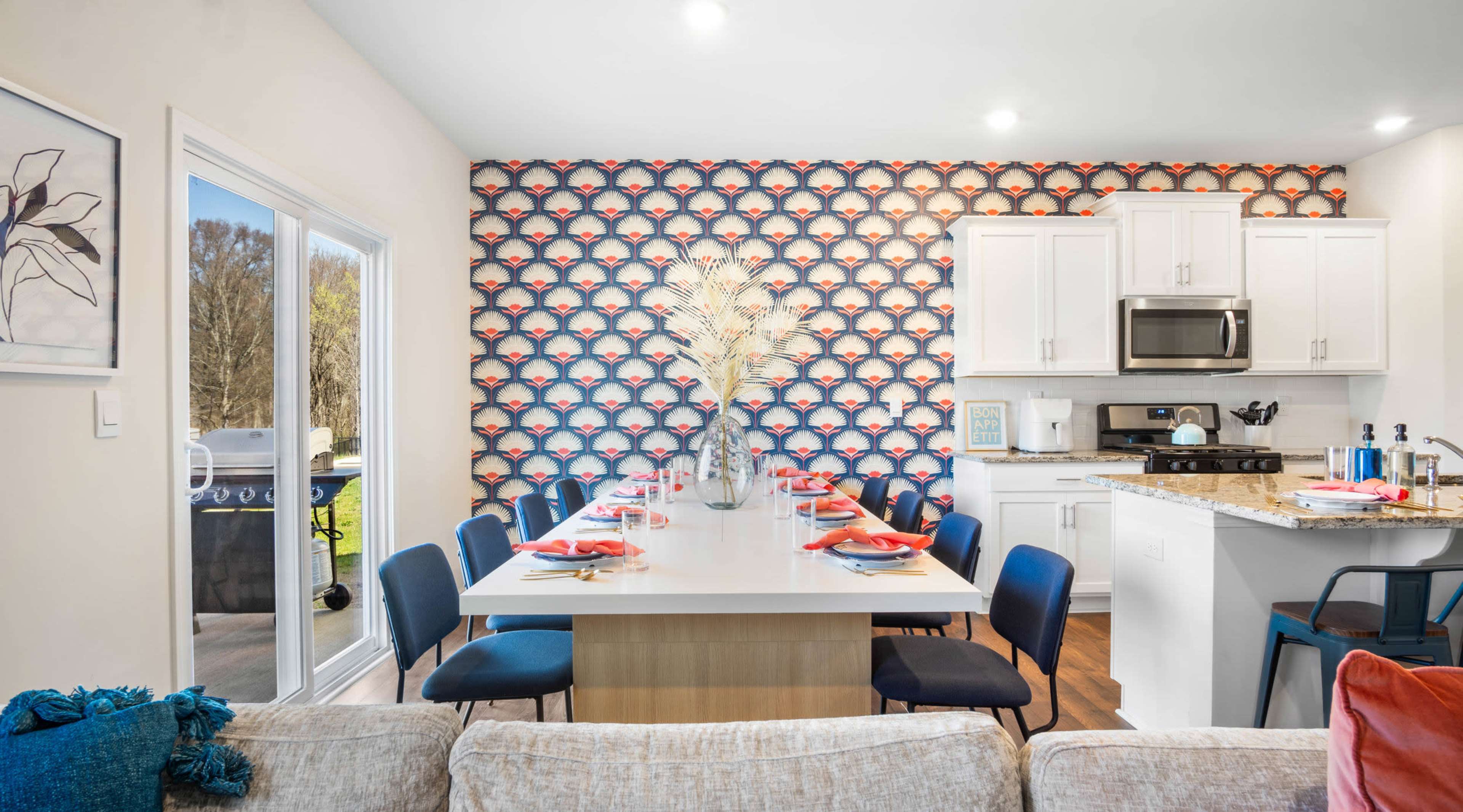 A modern dining area features a long table set for dinner against a colorful patterned accent wall, with an adjoining kitchen visible in the background.