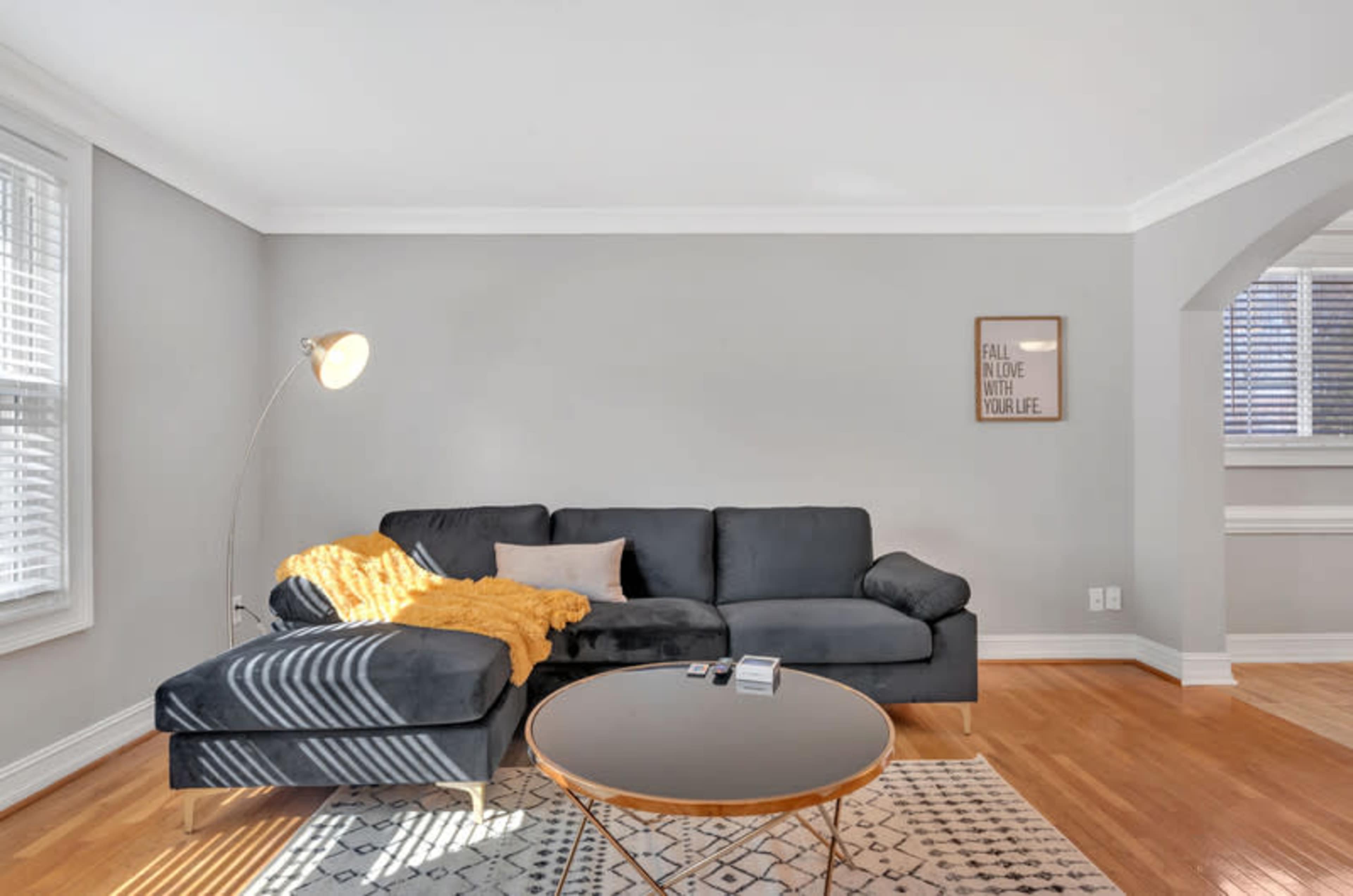 A modern living room features a dark sectional sofa with a yellow throw blanket, a round coffee table, and large windows allowing natural light to flow in.