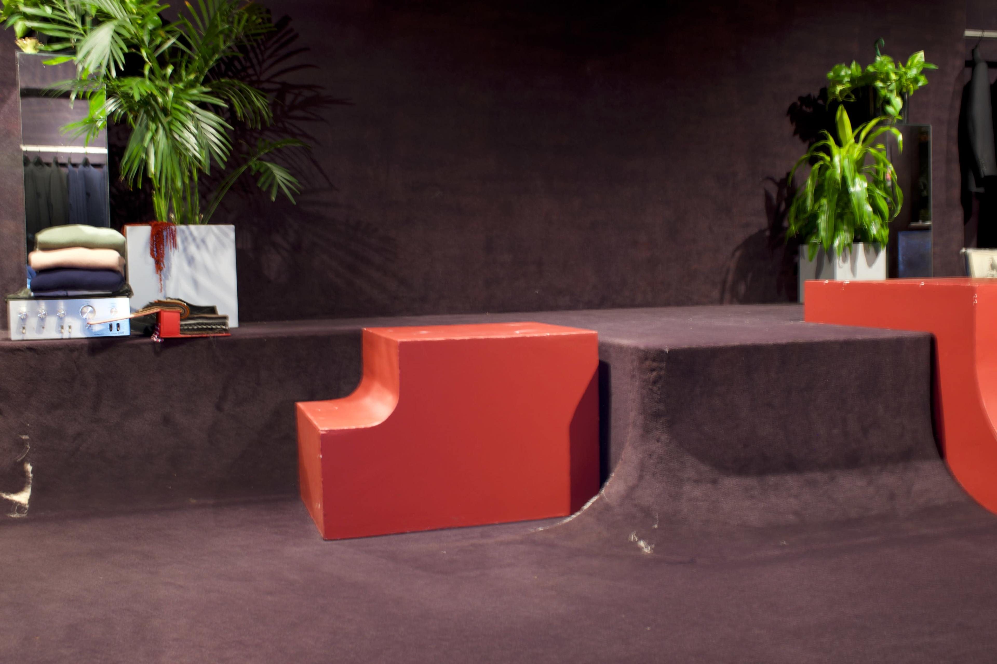 The scene features a red geometric object on a textured surface, flanked by potted plants and folded items in the background.