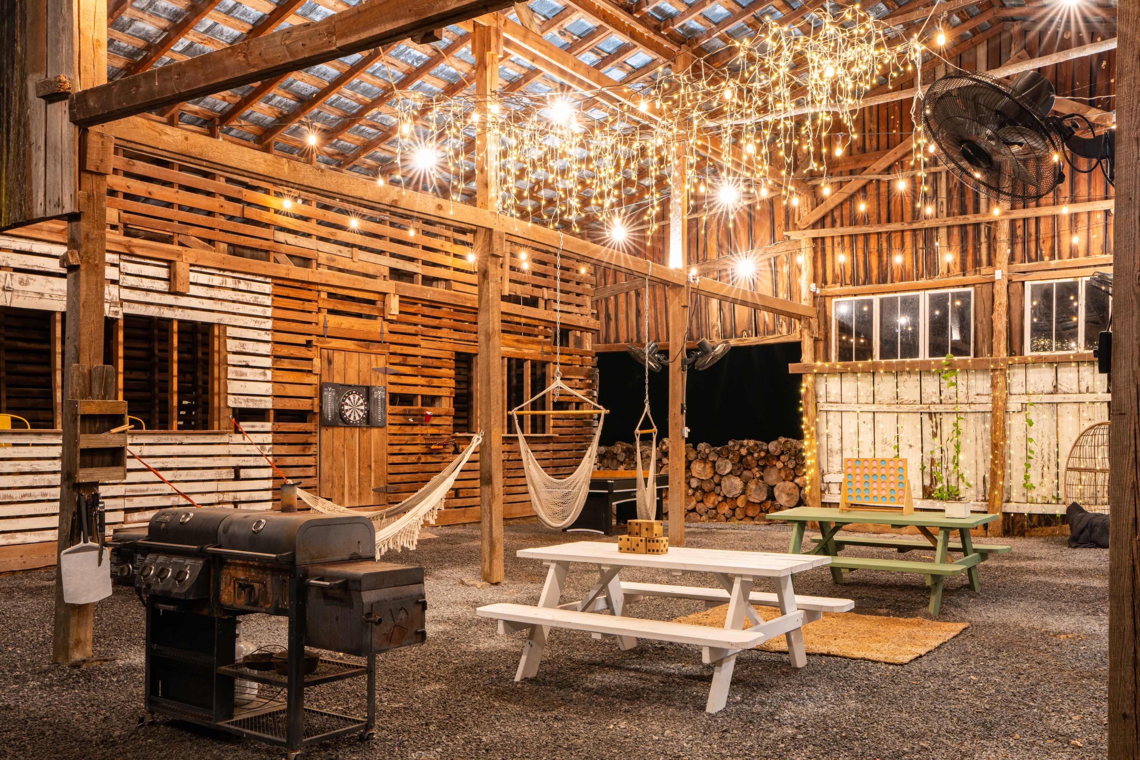The interior of a rustic barn features hanging lights, a picnic table, a grill, hammocks, and logs stacked in the background.