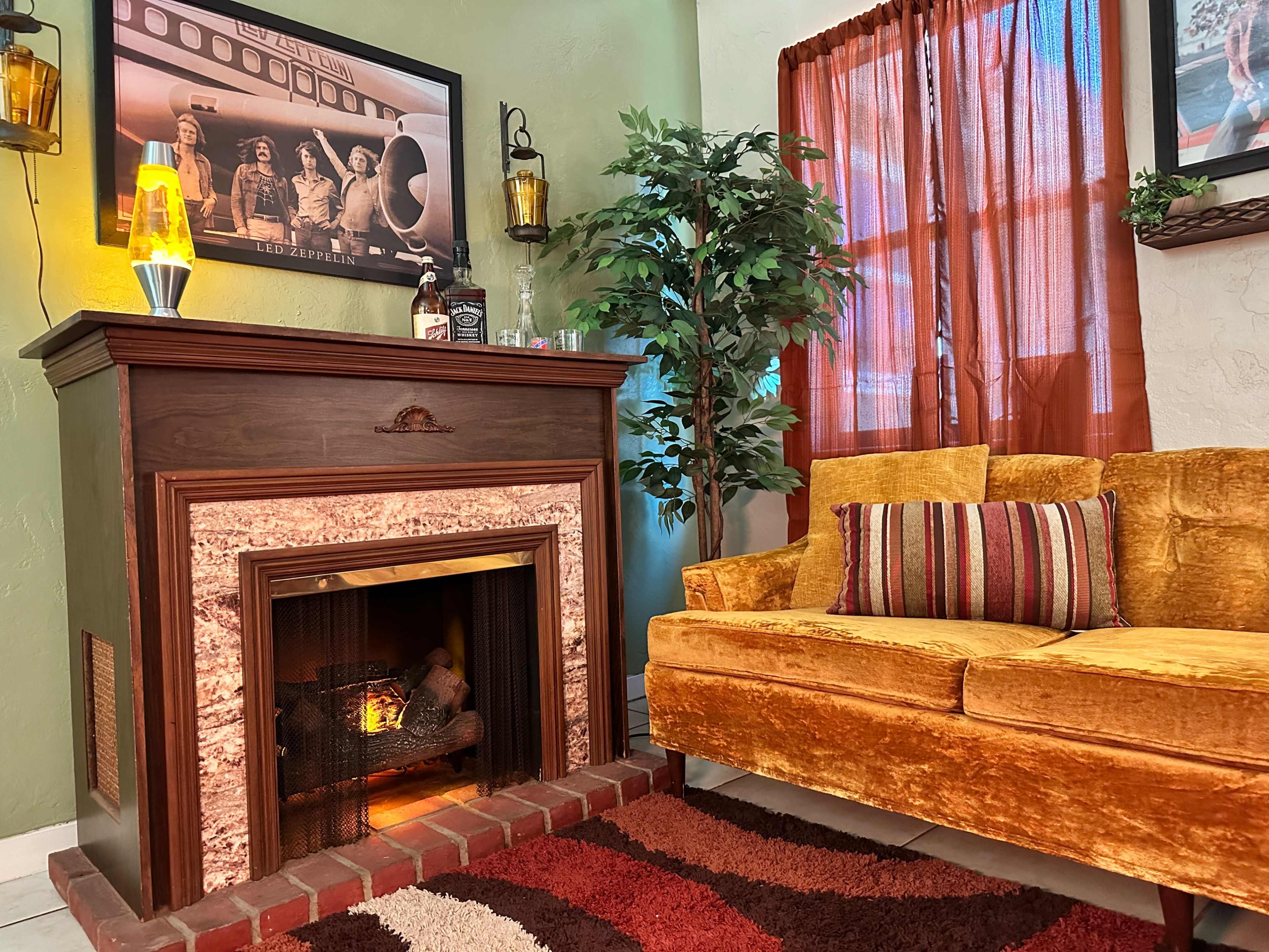 A cozy living room featuring a wooden fireplace, an orange velvet couch with decorative pillows, and a large window covered by a red curtain.