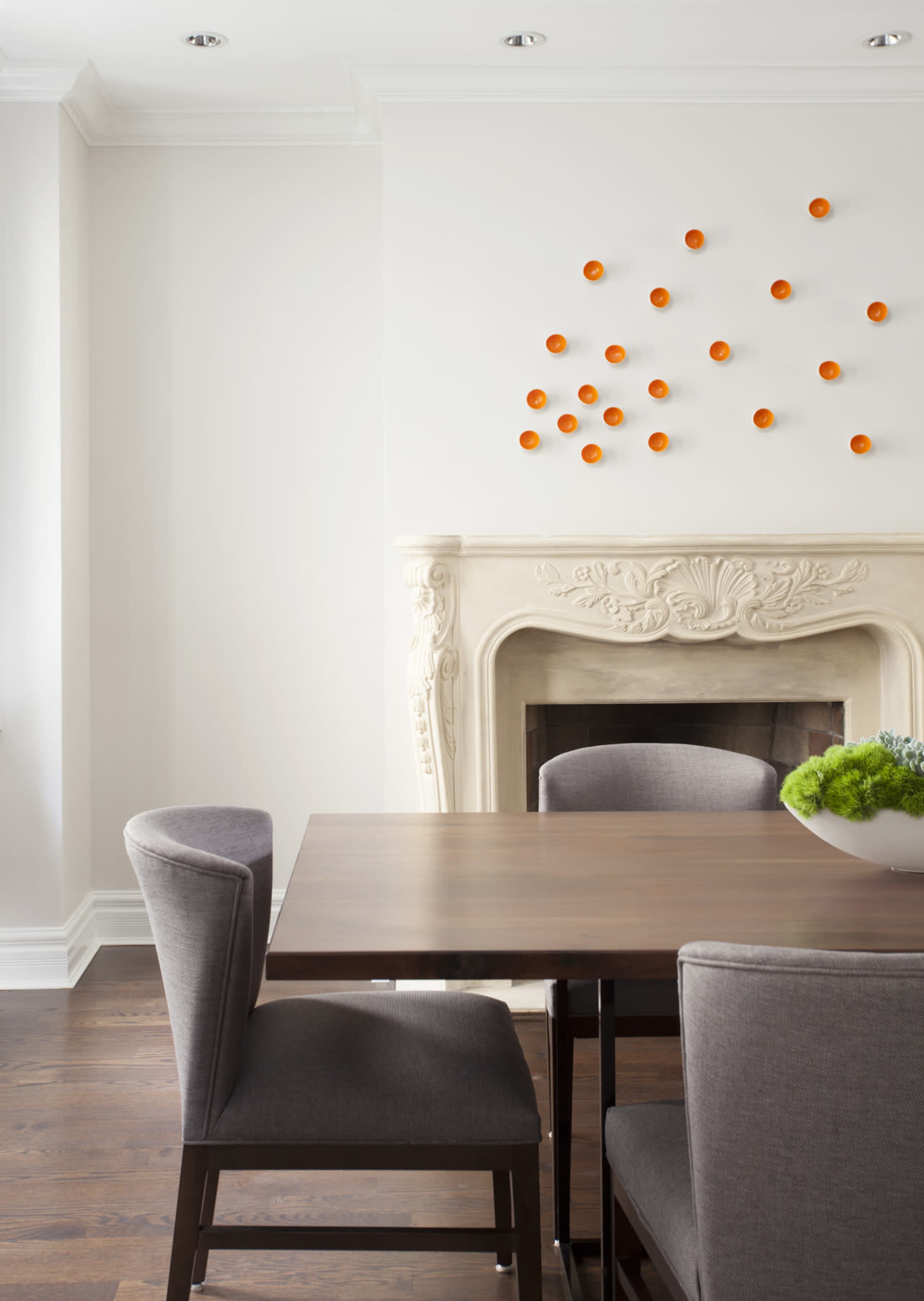 A modern dining area features a wooden table surrounded by gray chairs, with a decorative fireplace and orange wall art in the background.