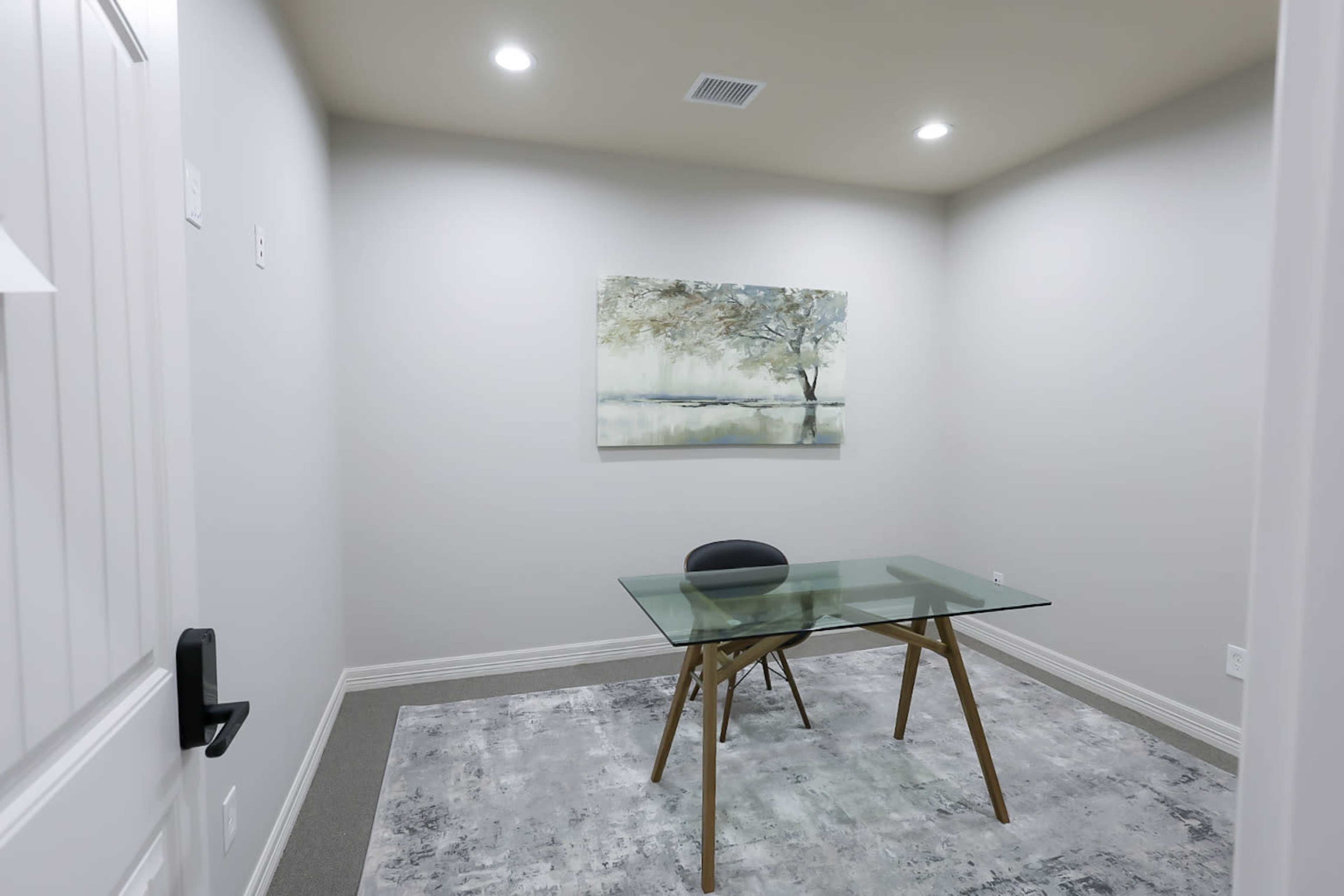 A small, brightly lit office space with a glass table, a black chair, and a landscape painting on the wall.