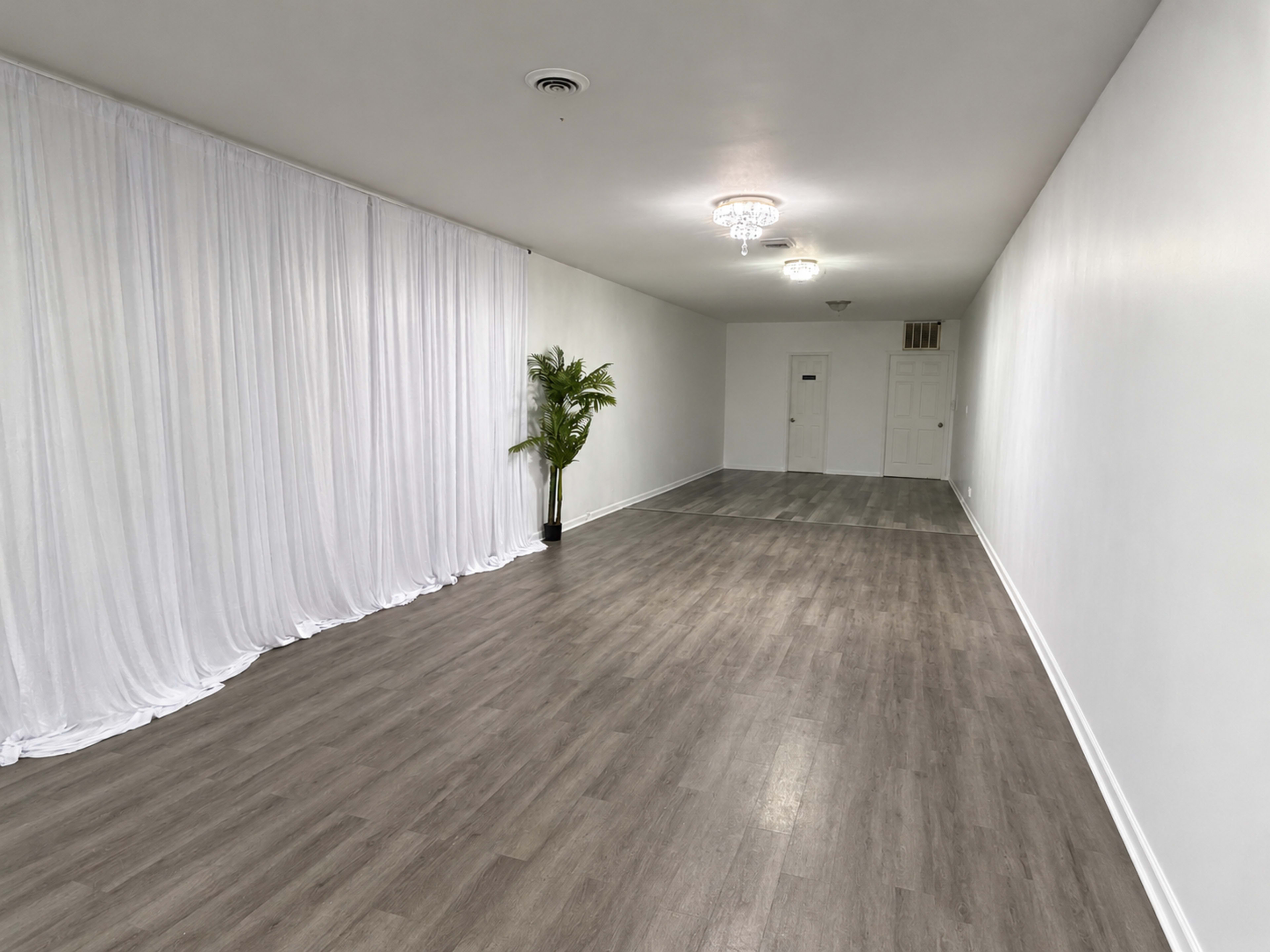 The image shows an empty, well-lit room with light wooden flooring, white walls, and a large white curtain along one side, complemented by a potted plant.