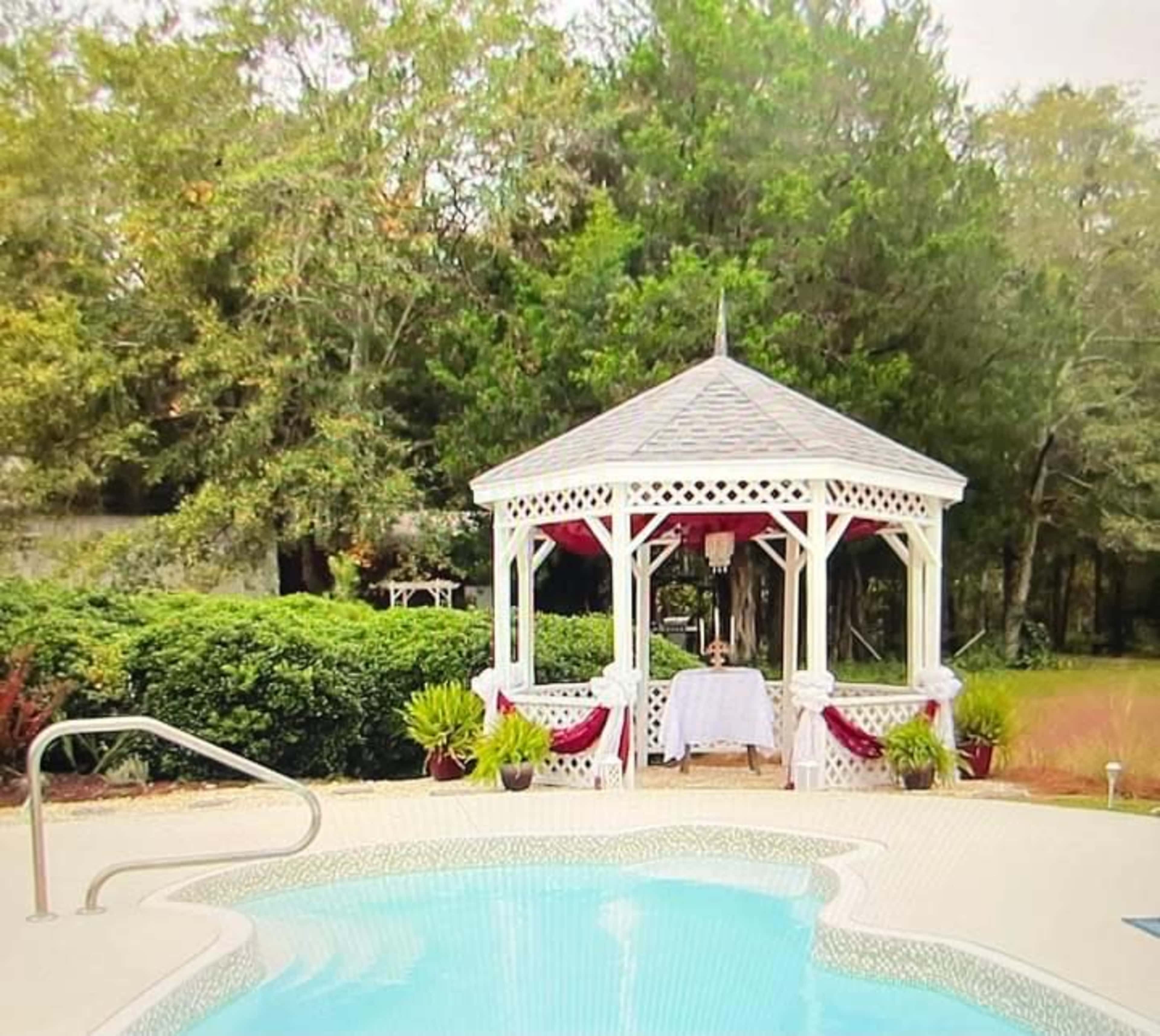 A white gazebo with red accents is situated near a sparkling pool, surrounded by greenery.