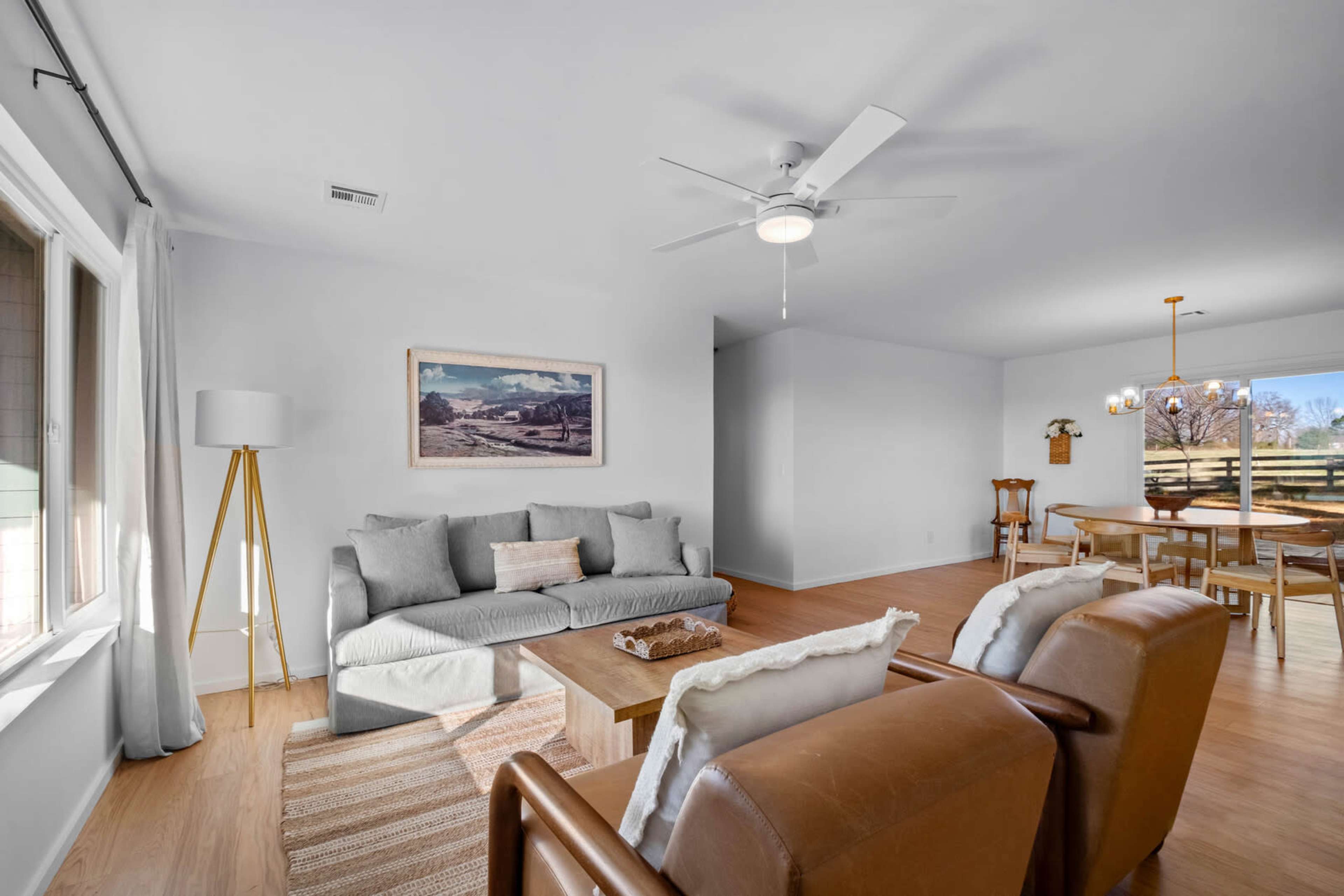 The image shows a bright, modern living room furnished with a gray sofa, brown armchairs, and a wooden dining area in the background.