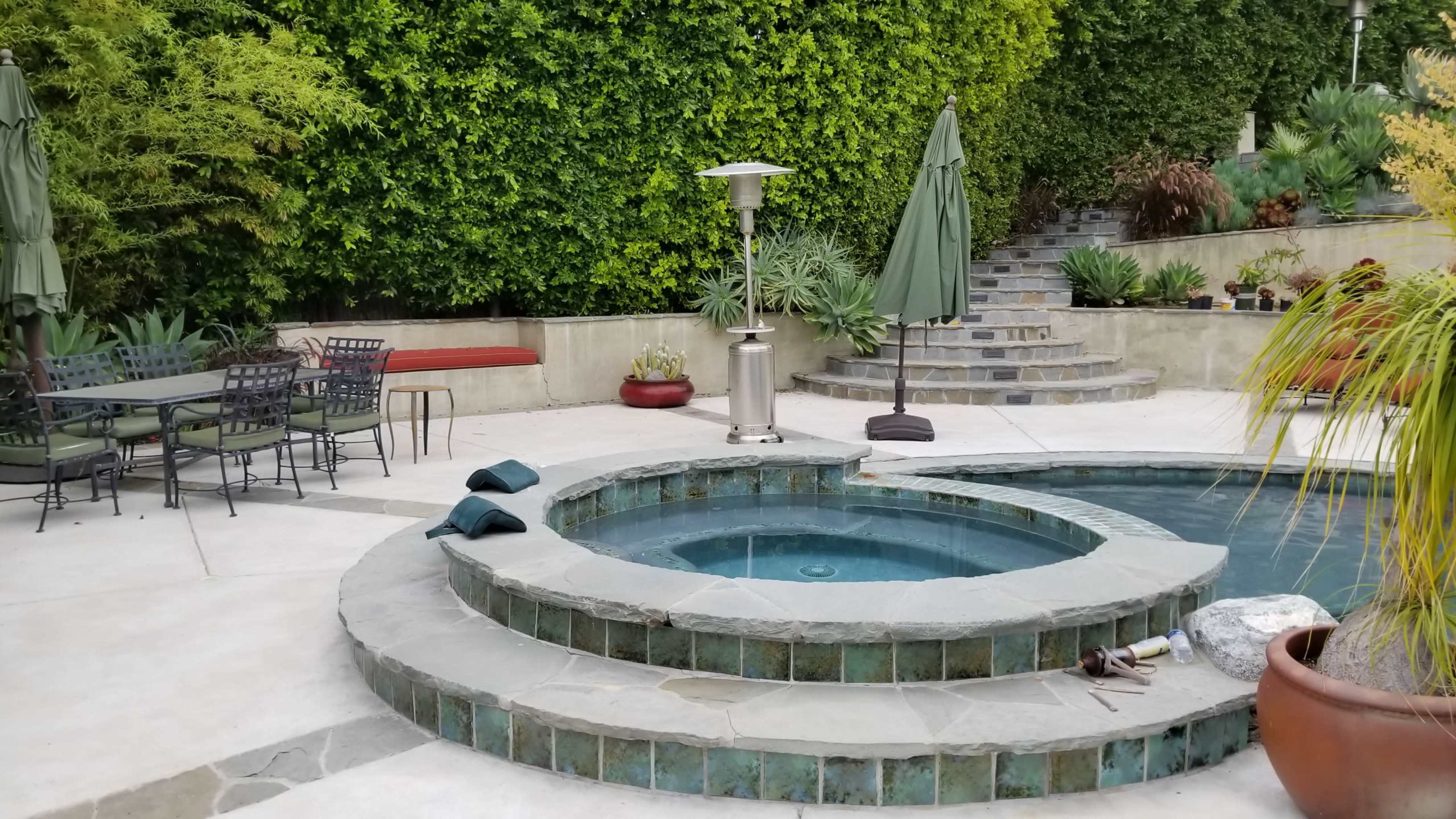 A landscaped patio area with a spa, heating lamp, outdoor seating, and lush greenery in the background.