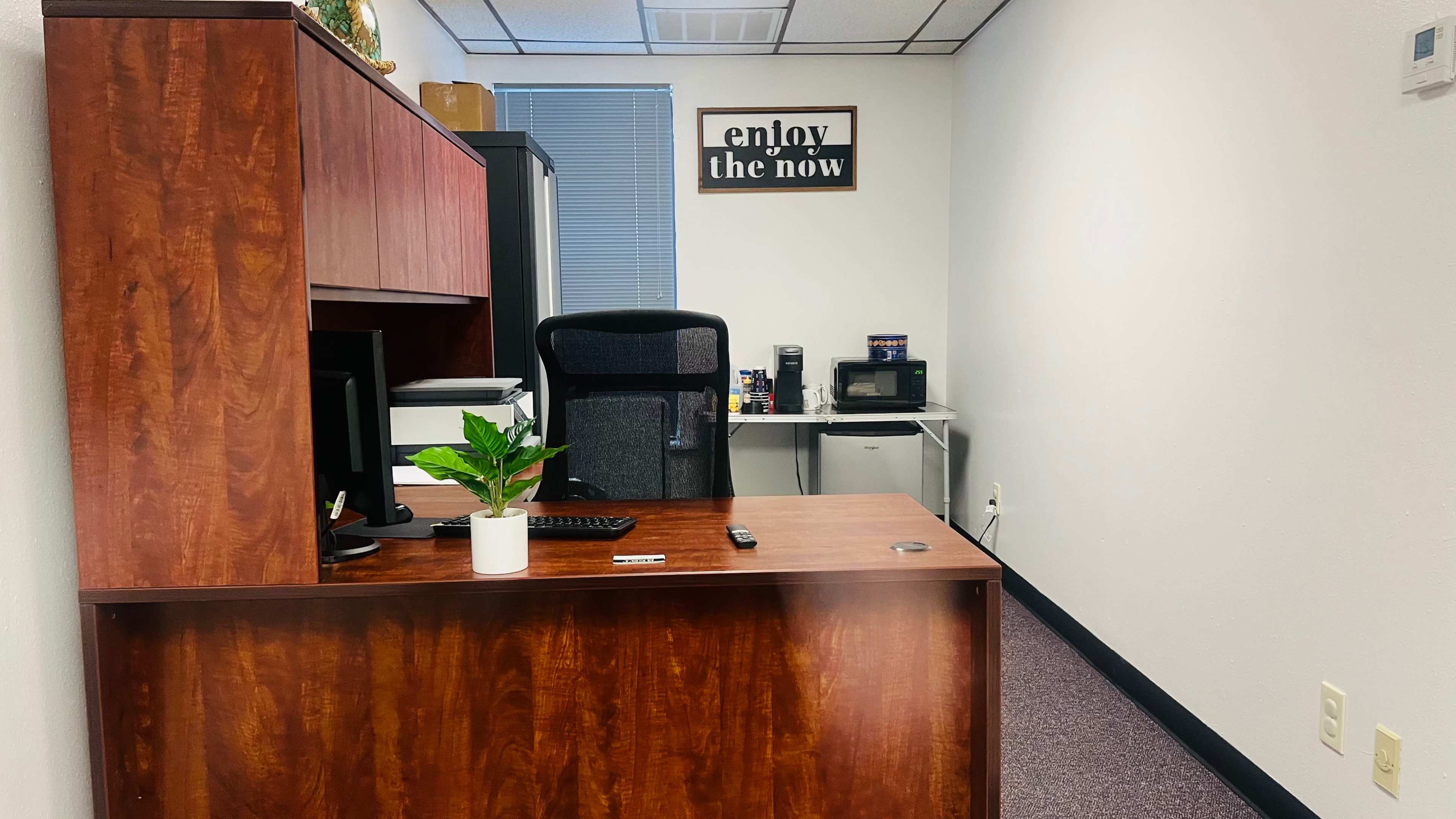 An office space features a wooden desk with a computer, a chair, and a white plant pot, along with a microwave and cabinets in the background.