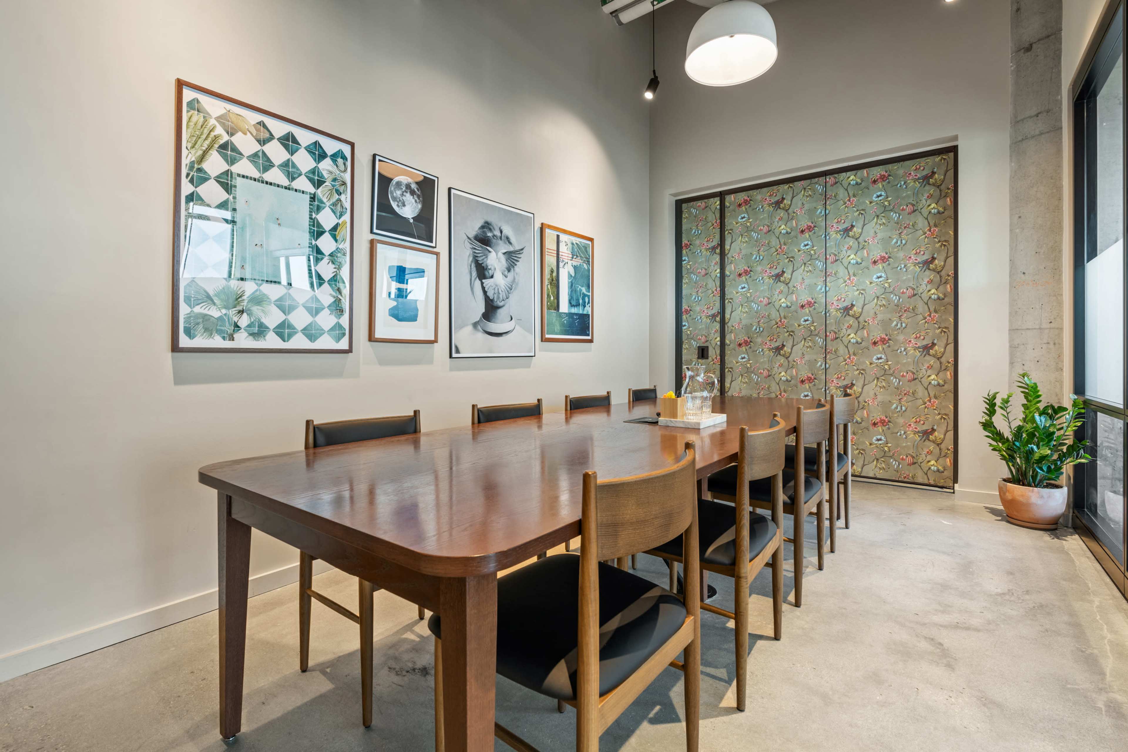 A long wooden table with ten chairs is set in a well-lit room adorned with framed artworks and a sliding door featuring floral patterns.