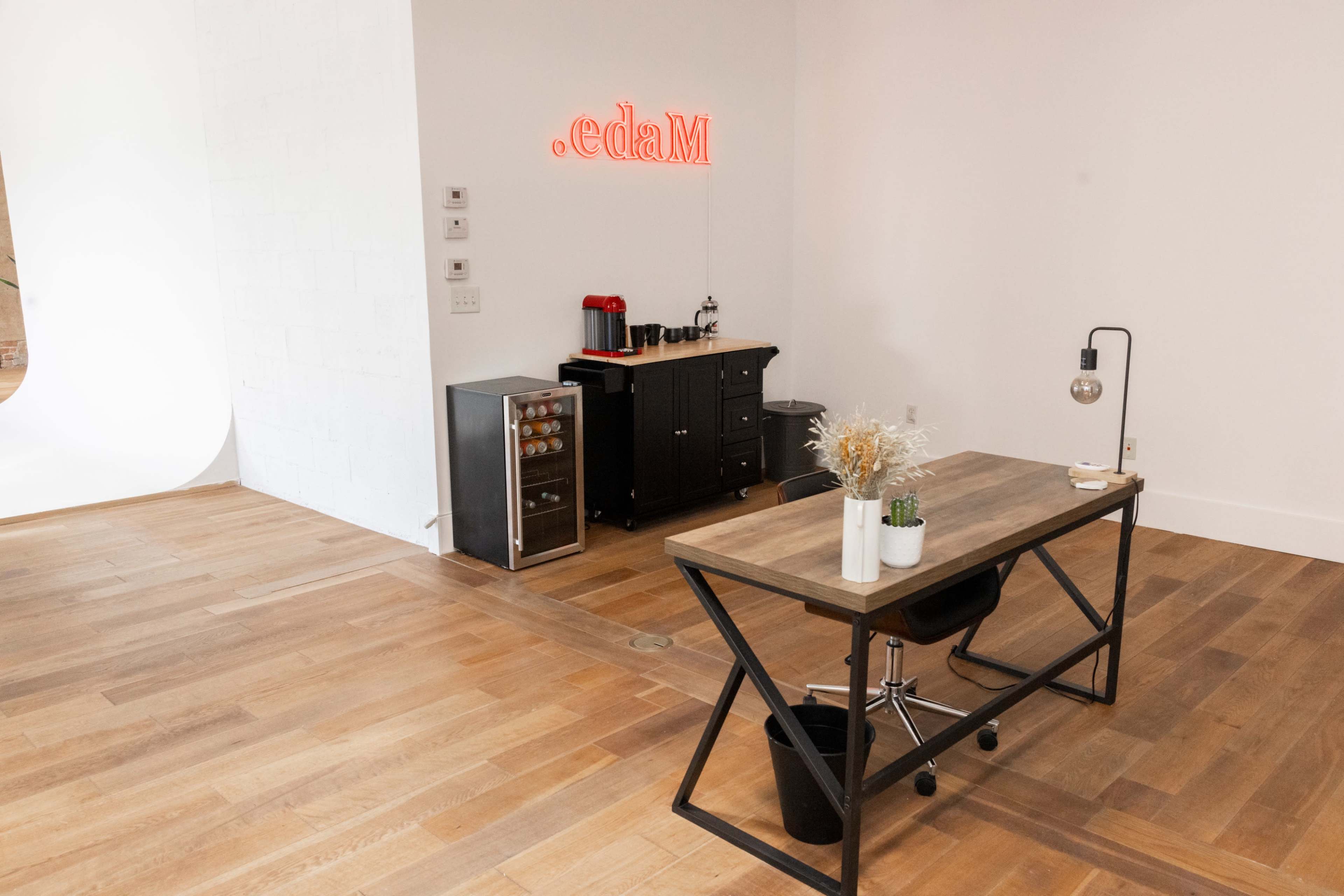 A modern office space features a wooden desk, a coffee station, a small refrigerator, and a wall with a neon sign.