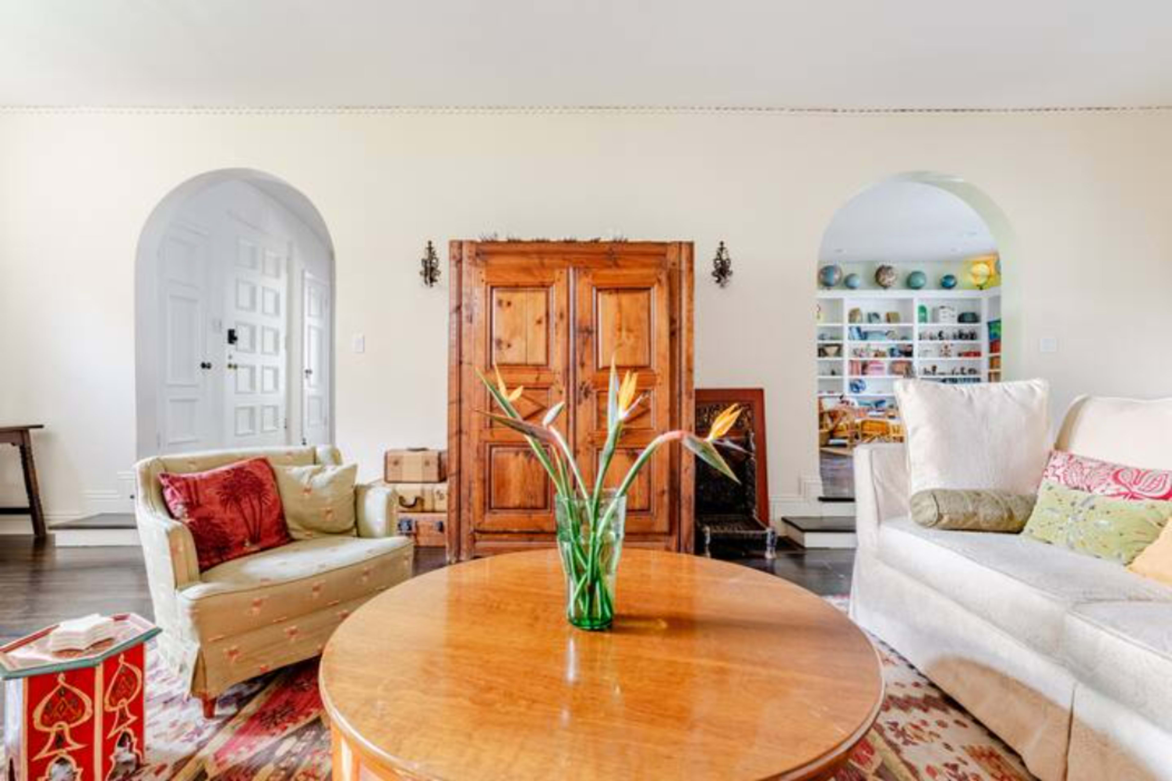 The image shows a living room with a round wooden coffee table, two sofas, and an arched doorway leading to a shelf filled with colorful items.