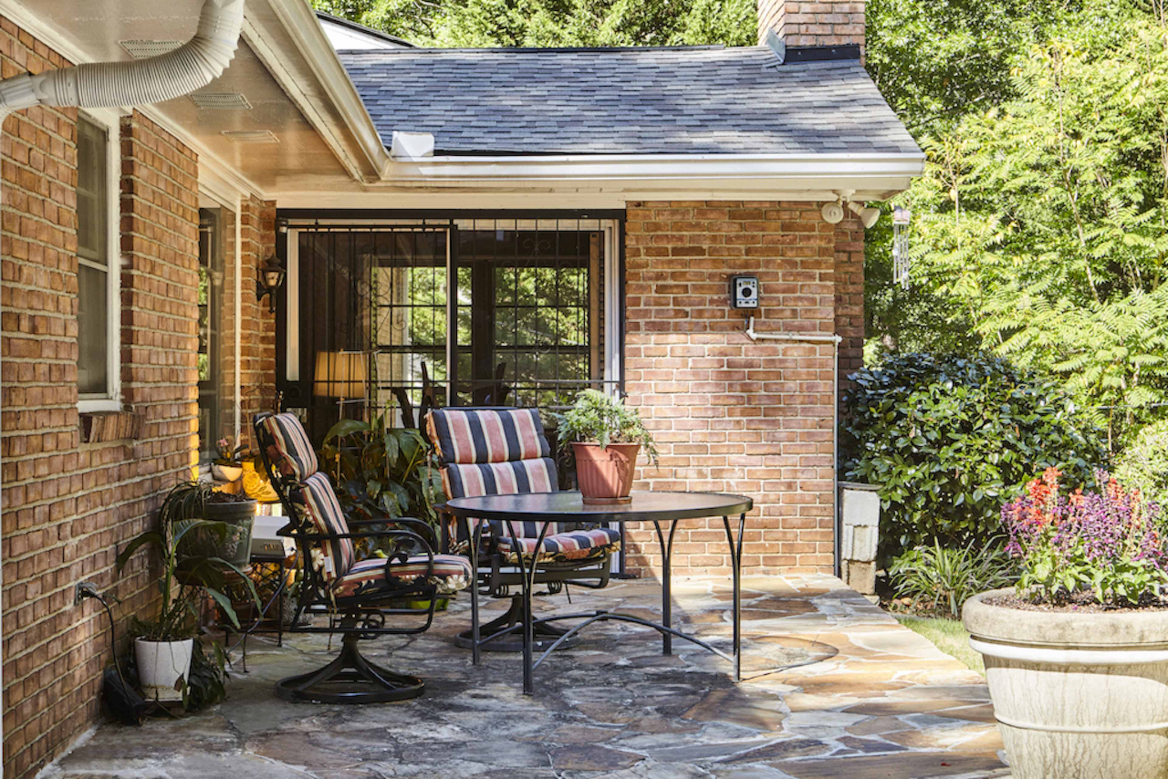 A patio area features a round table with striped chairs, surrounded by greenery and blooming flowers, attached to a brick house.