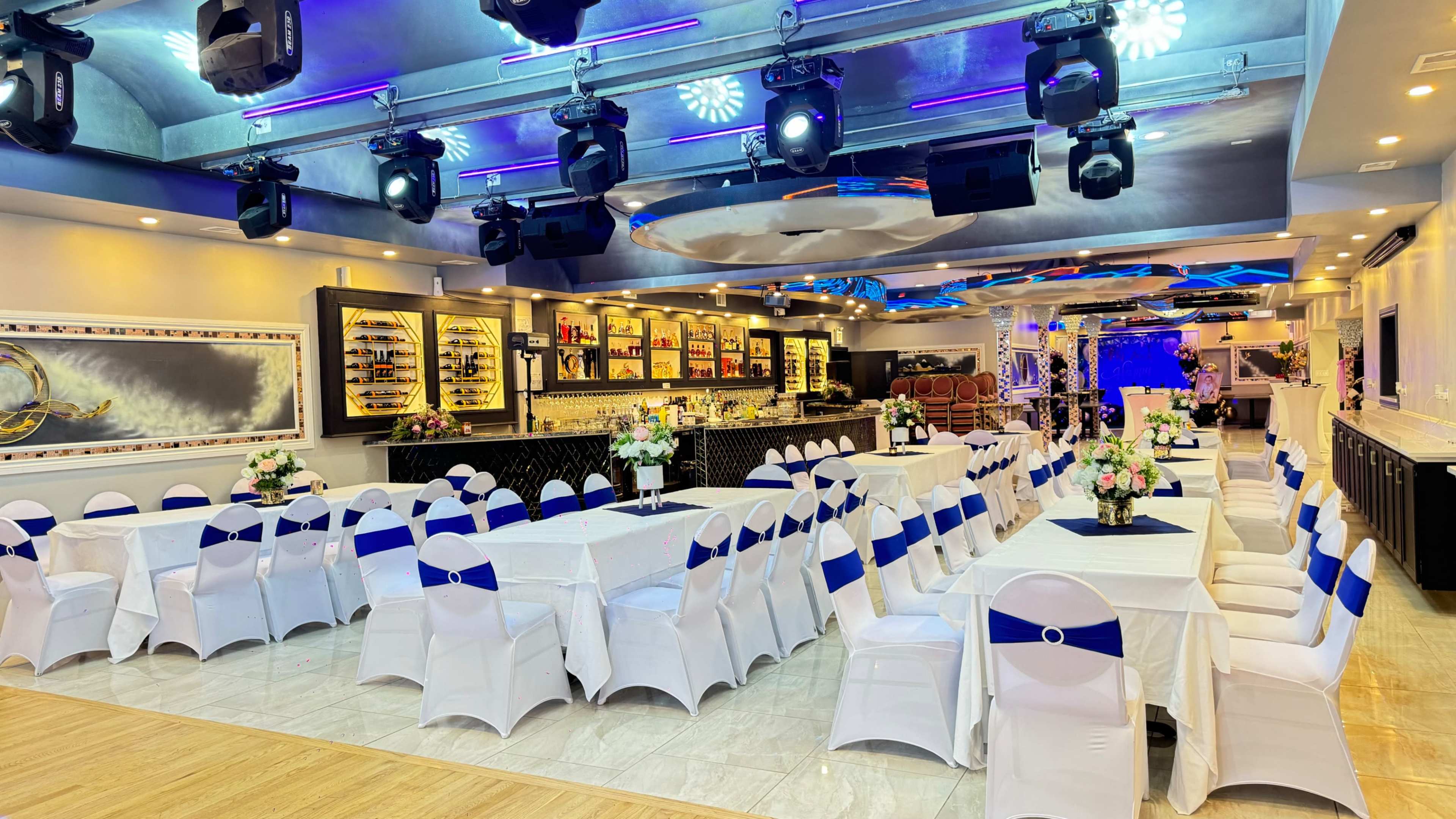 The image shows a spacious event hall set up with several tables covered in white linens and blue sashes, with floral centerpieces and modern lighting fixtures.