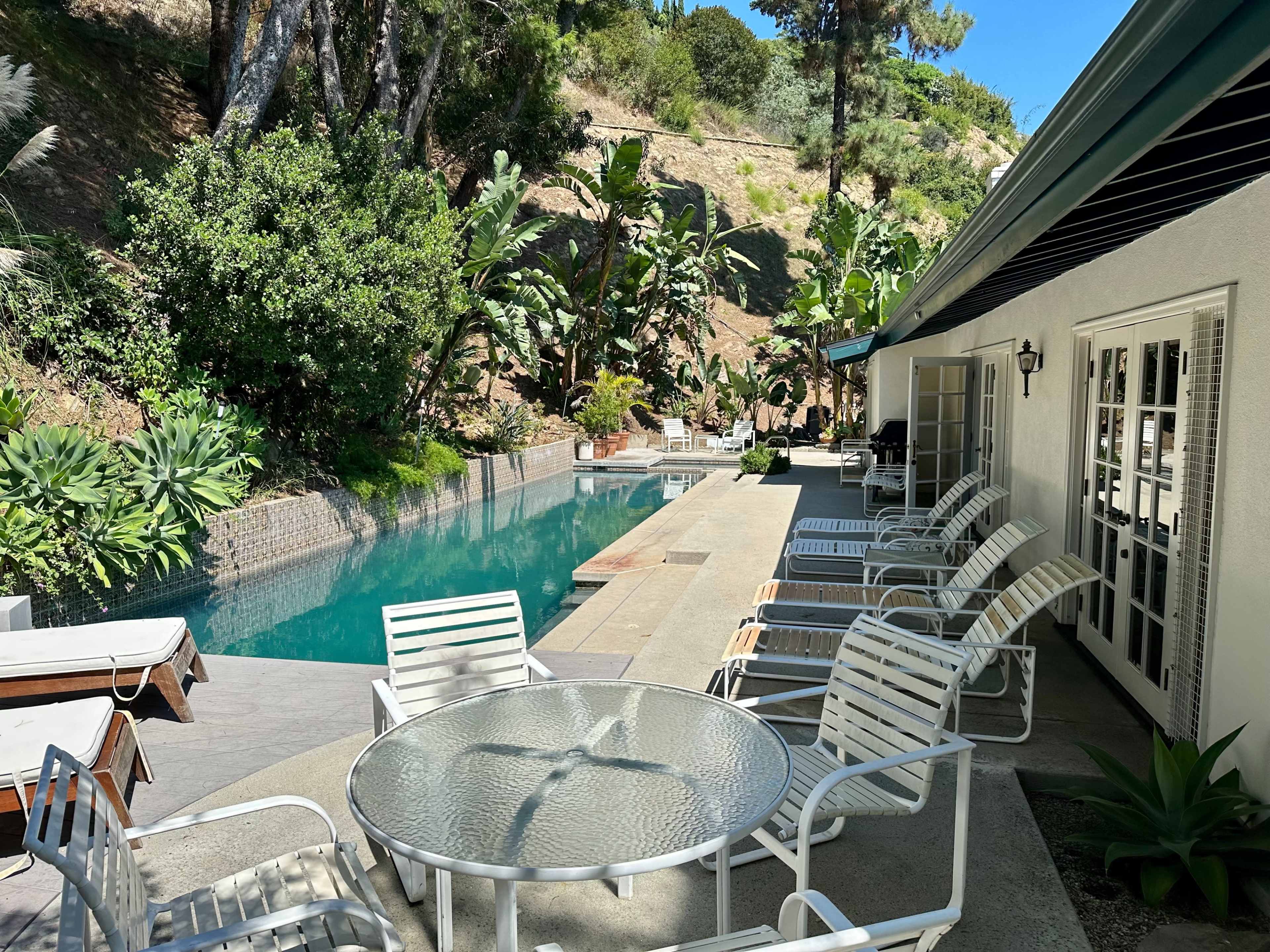 A backyard features a swimming pool lined with lounge chairs and surrounded by lush greenery.