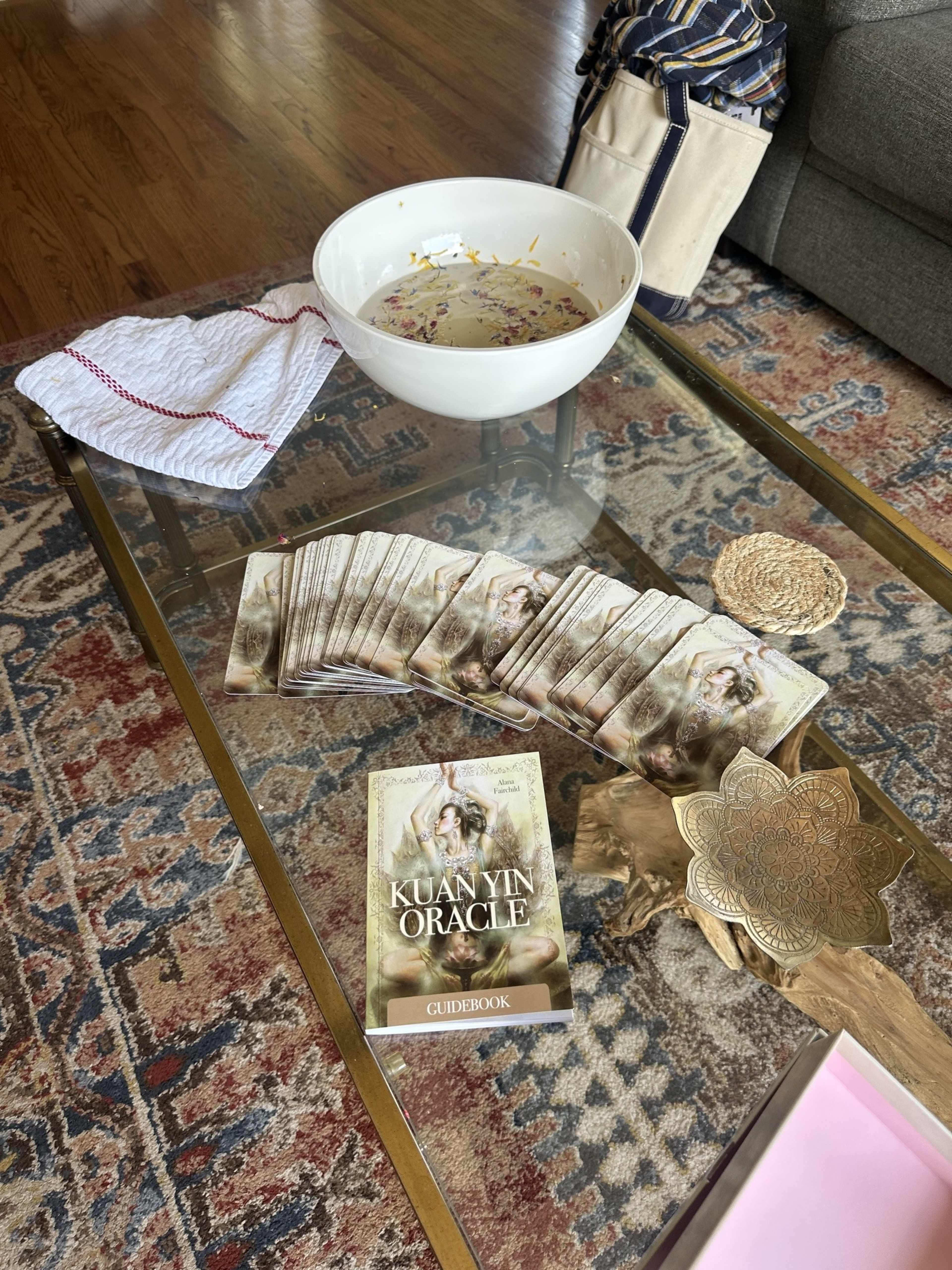 A coffee table is set with a bowl, a folded towel, a decorative plate, and a stack of Kuan Yin Oracle cards alongside their guidebook.