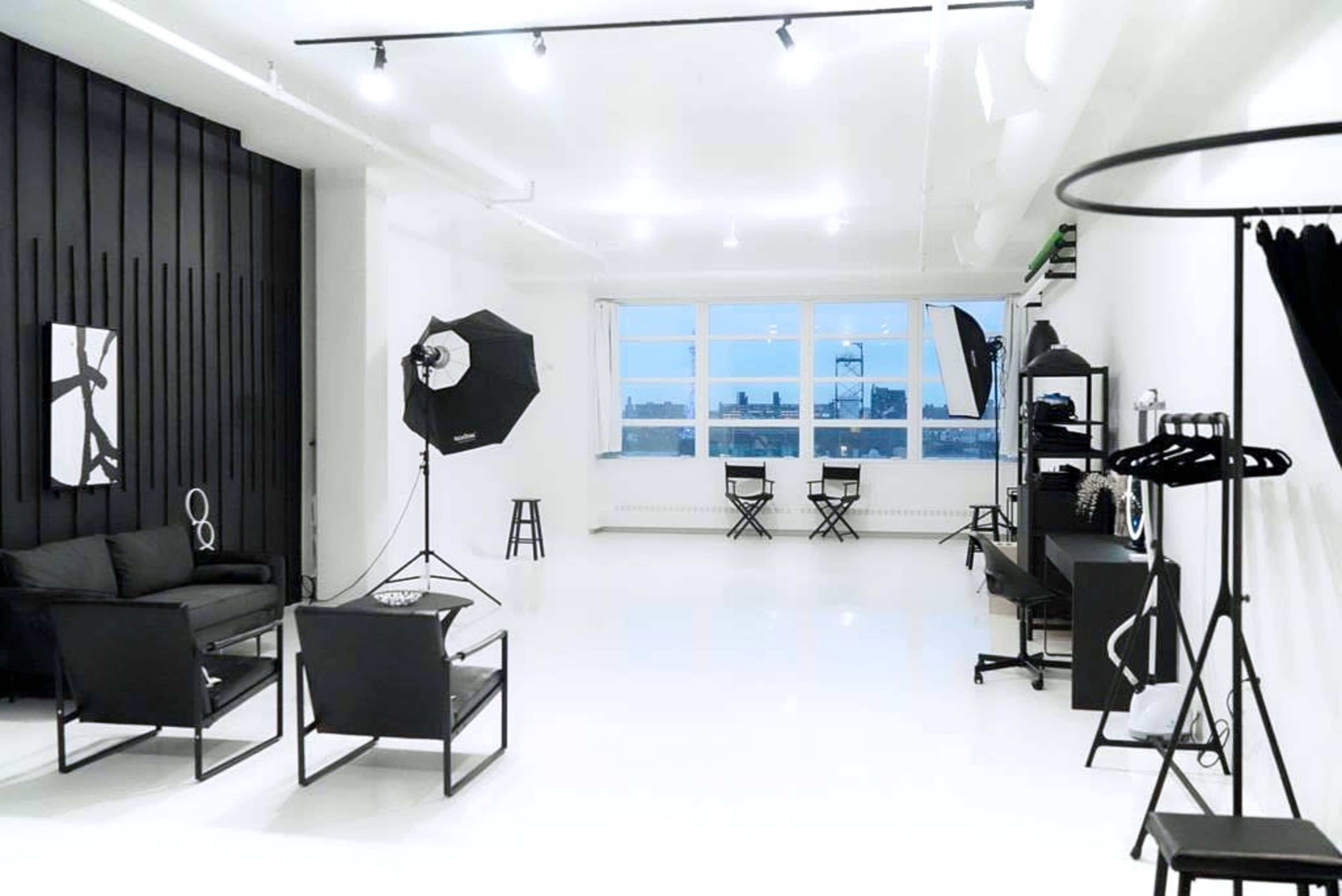 A spacious, well-lit studio featuring black furniture, a photography backdrop, and two makeup chairs near large windows.