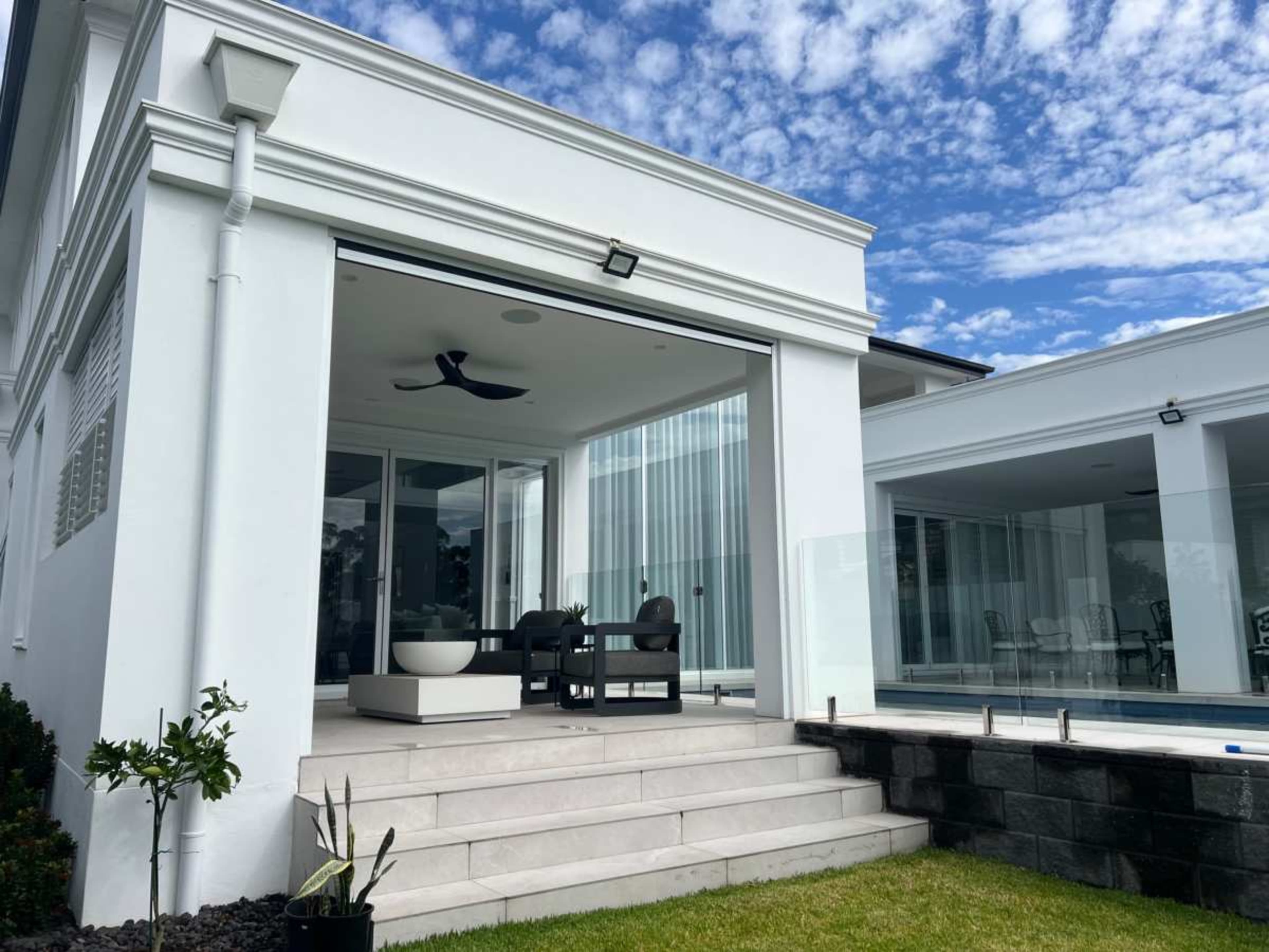 The image shows a modern white house with large glass windows, a patio area with seating, and a neatly maintained lawn.
