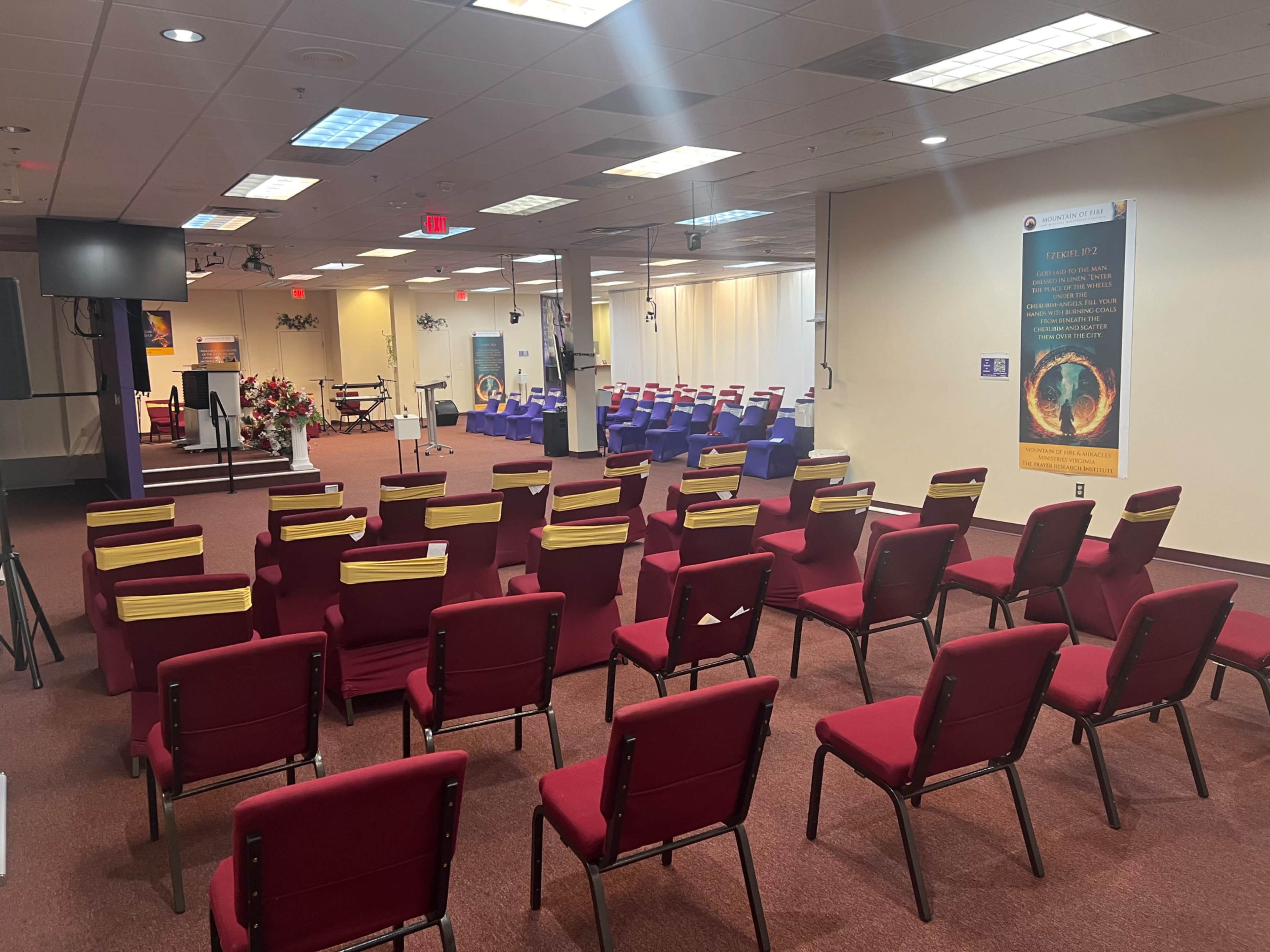 The image shows a spacious indoor venue with rows of red chairs facing a stage area, decorated with plants and a projector screen.