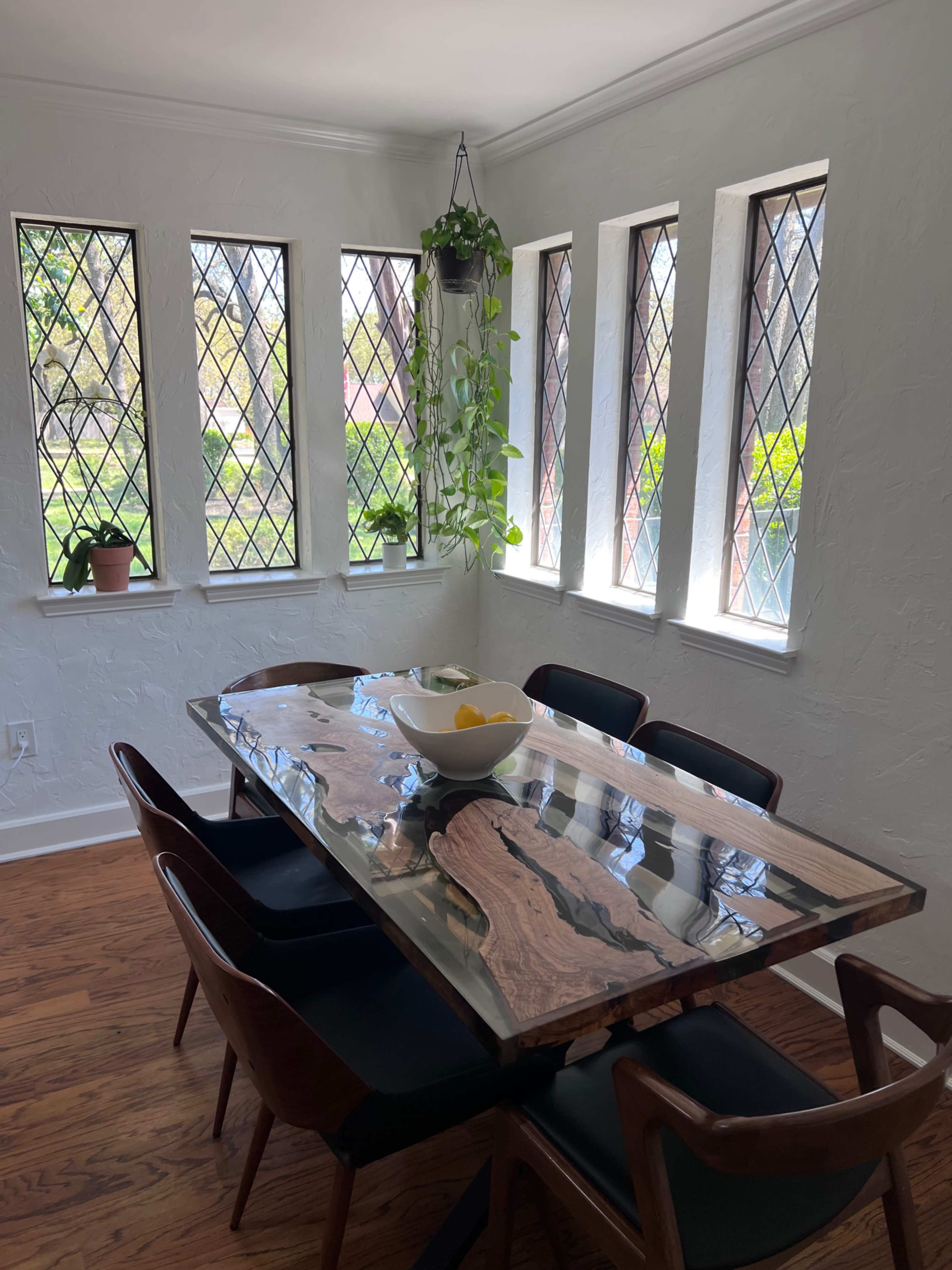 A dining area with a wooden table and eight chairs, surrounded by windows with diamond-style panes and plants.