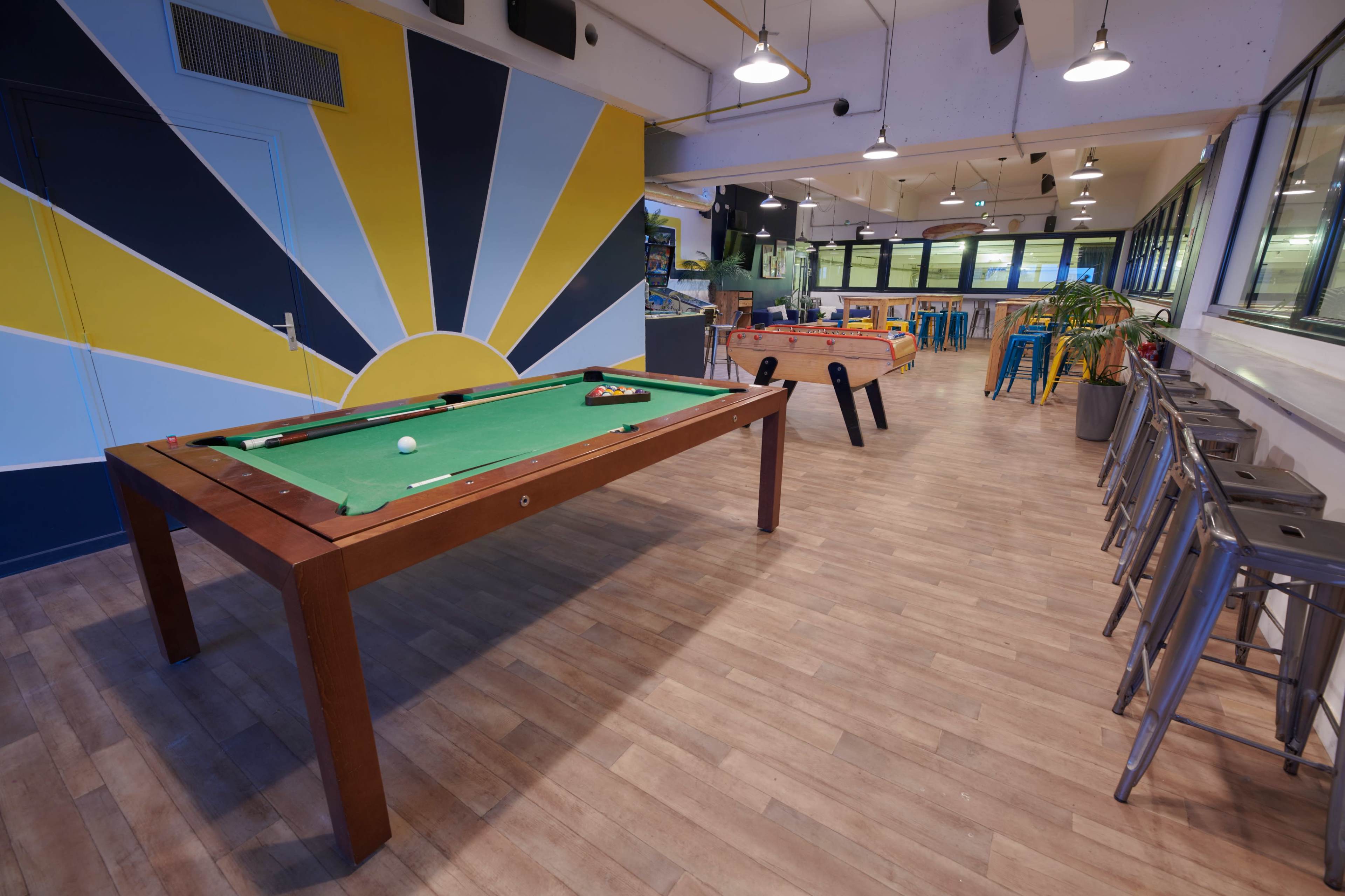A recreational room with a green billiard table, colorful wall art, and seating arrangements, alongside foosball and dining tables.