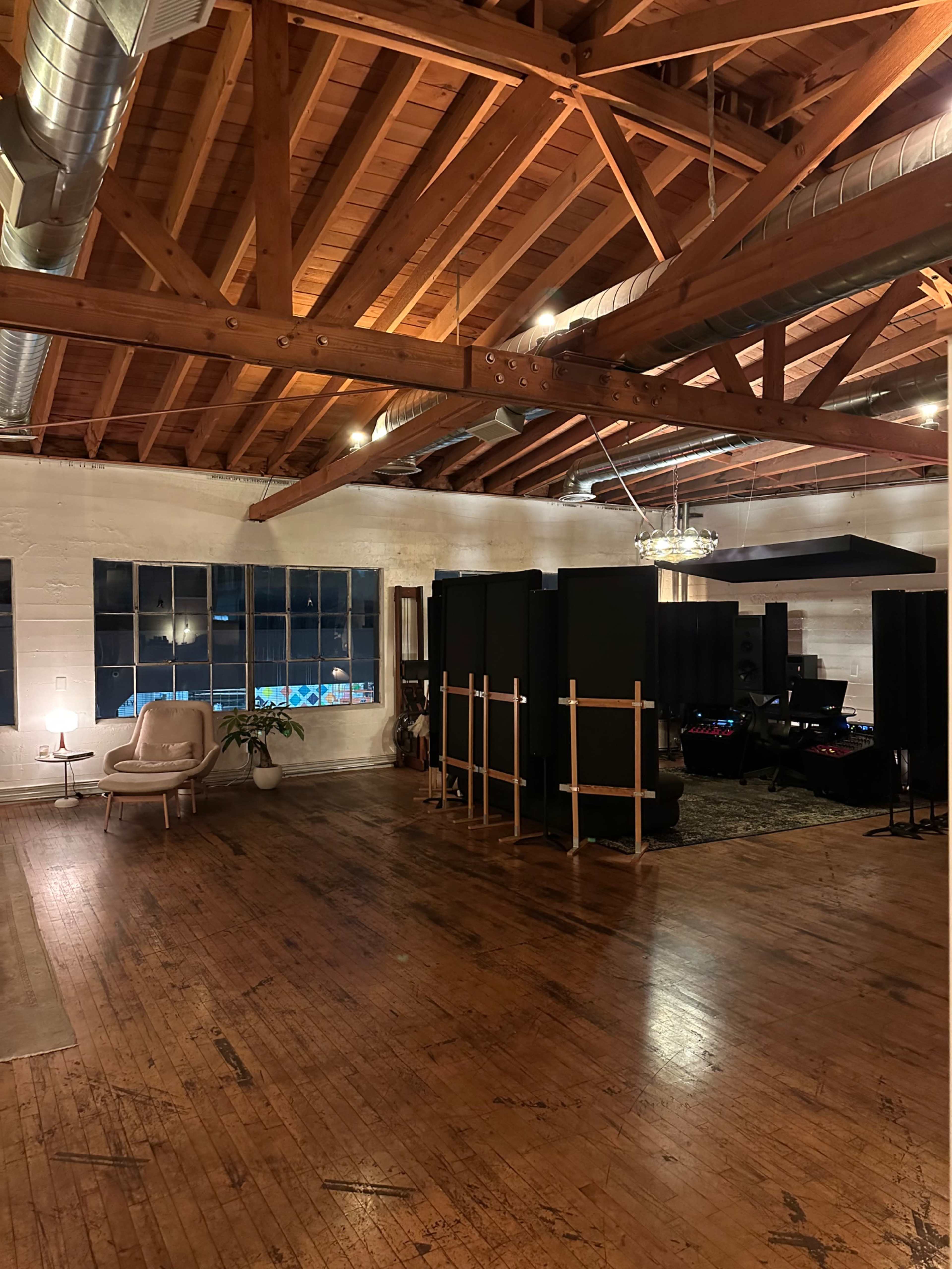 A spacious studio or workspace with exposed wooden beams, concrete walls, and a seating area featuring a chair, alongside sound equipment partially obscured by black dividers.