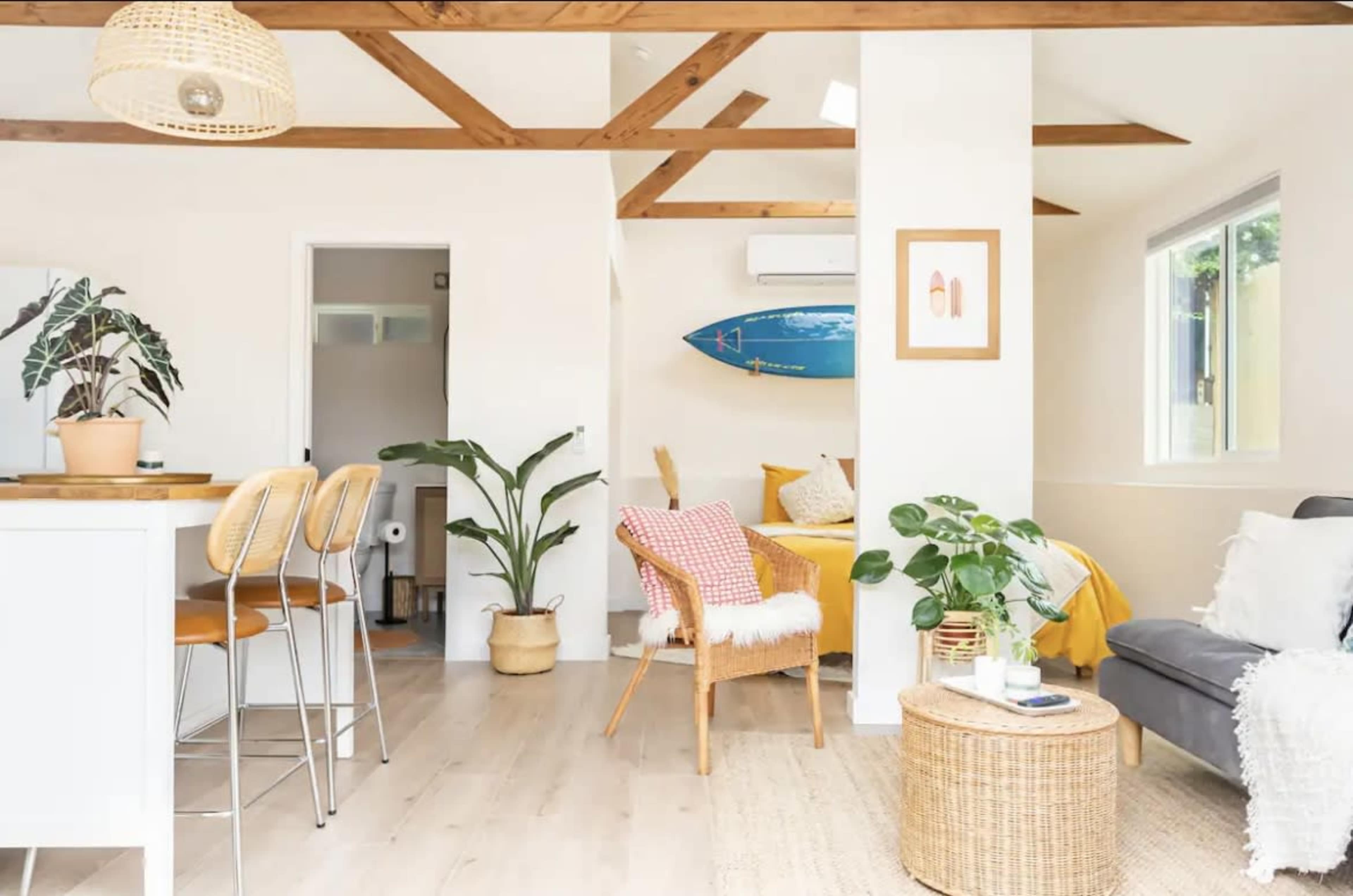 The image shows a bright, open living space with wooden beams, a kitchen island, a cozy seating area, and a surfboard on the wall.