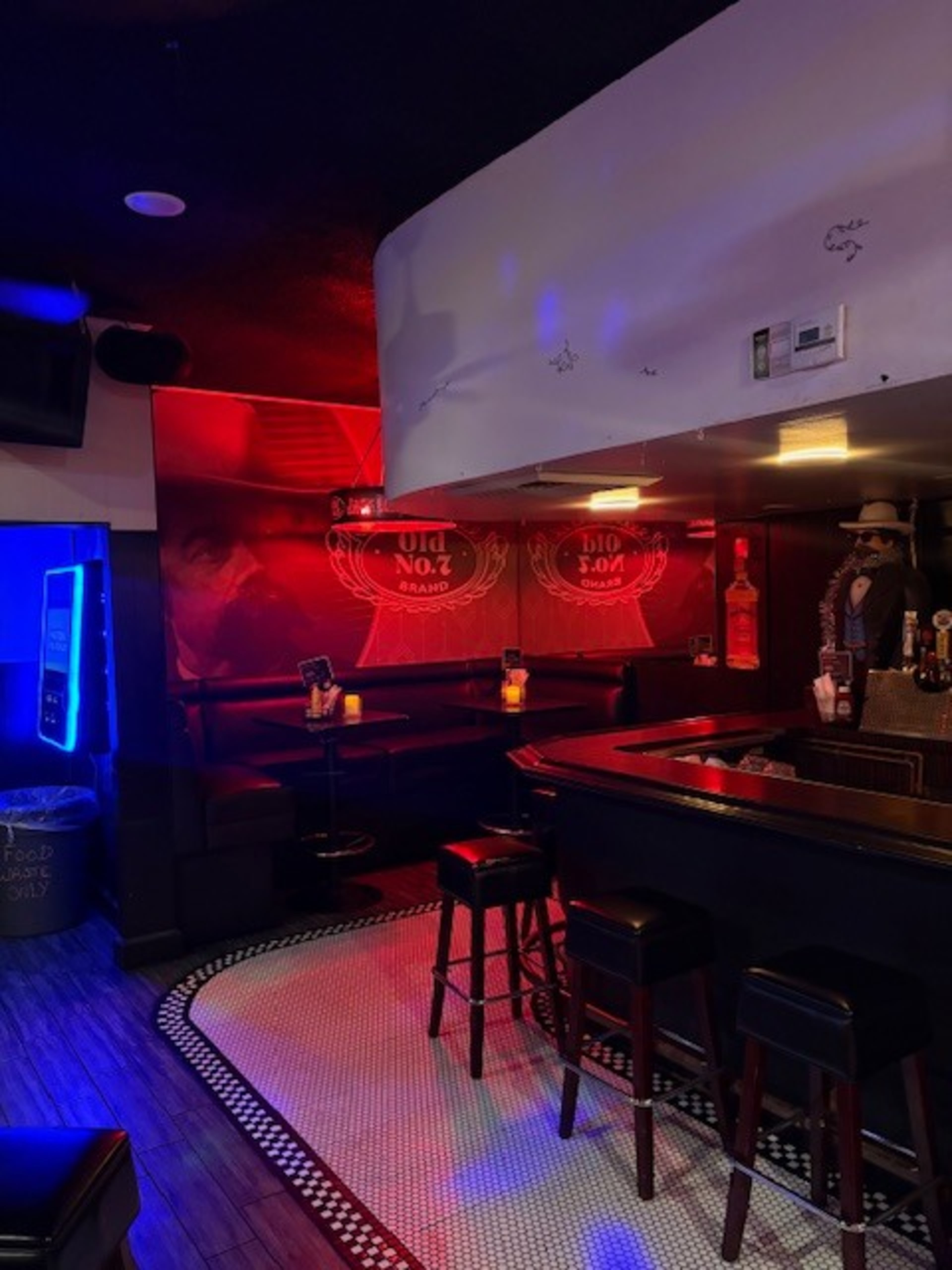 The image shows the interior of a bar featuring a dimly lit space with red and blue lighting, a wooden bar with stools, and a patterned floor.