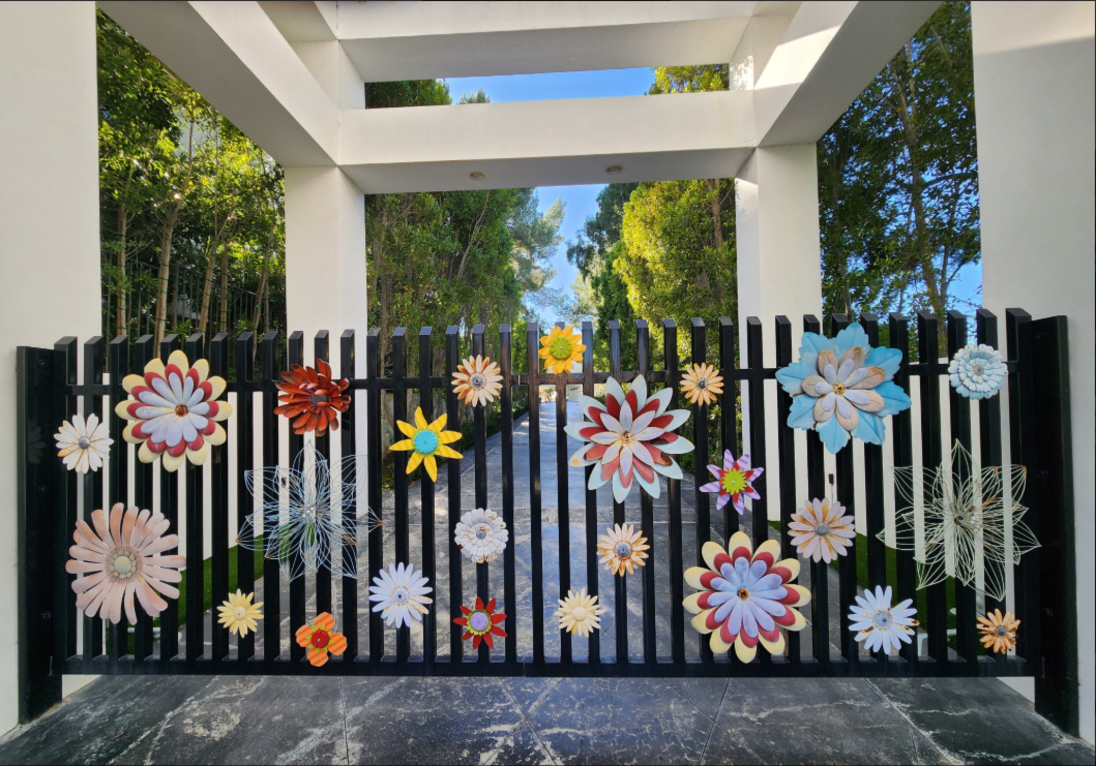 A decorative black fence adorned with various colorful flower designs stands in a landscaped area with trees in the background.