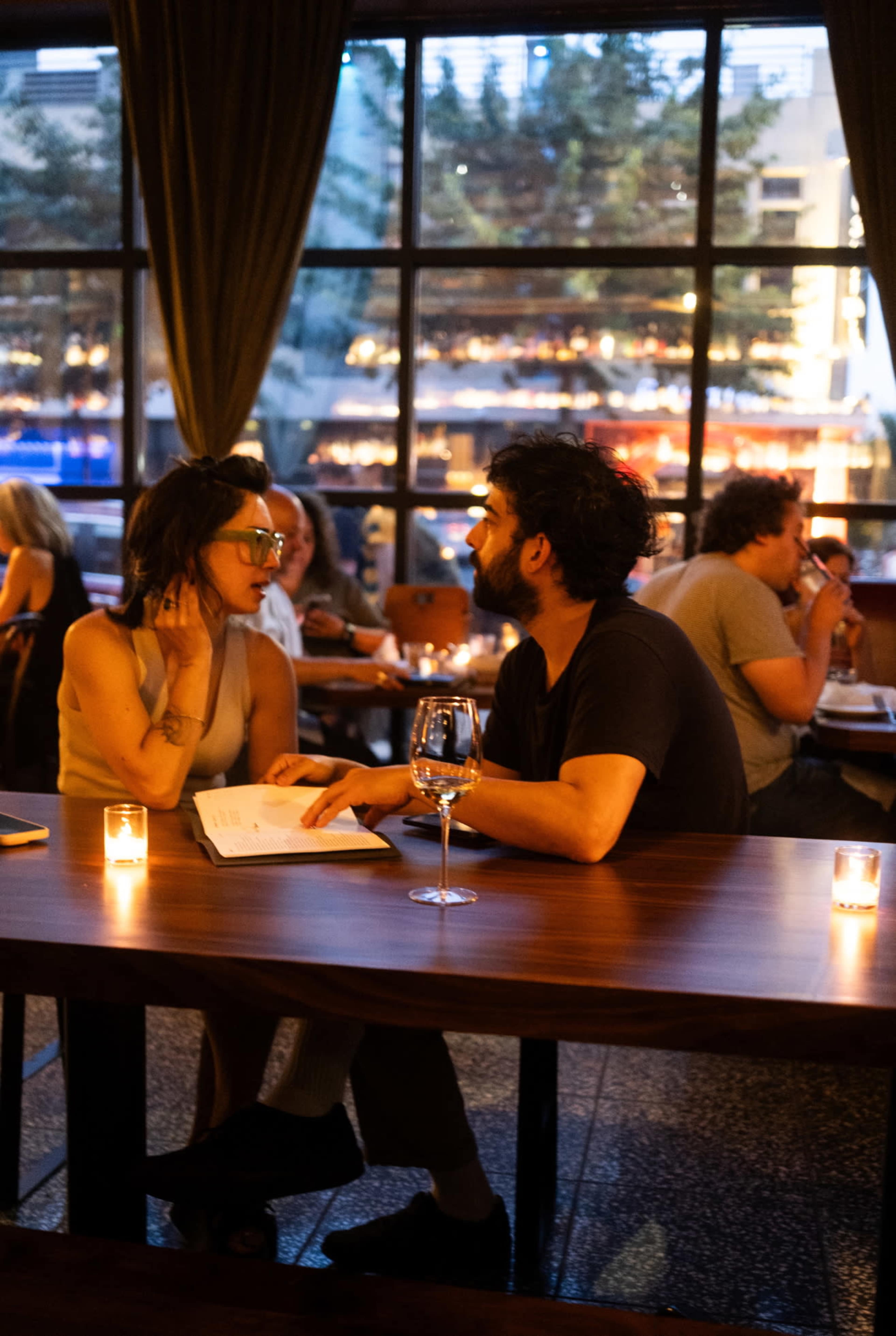 A couple sits at a dimly lit table in a restaurant, looking at a menu while other patrons dine nearby.